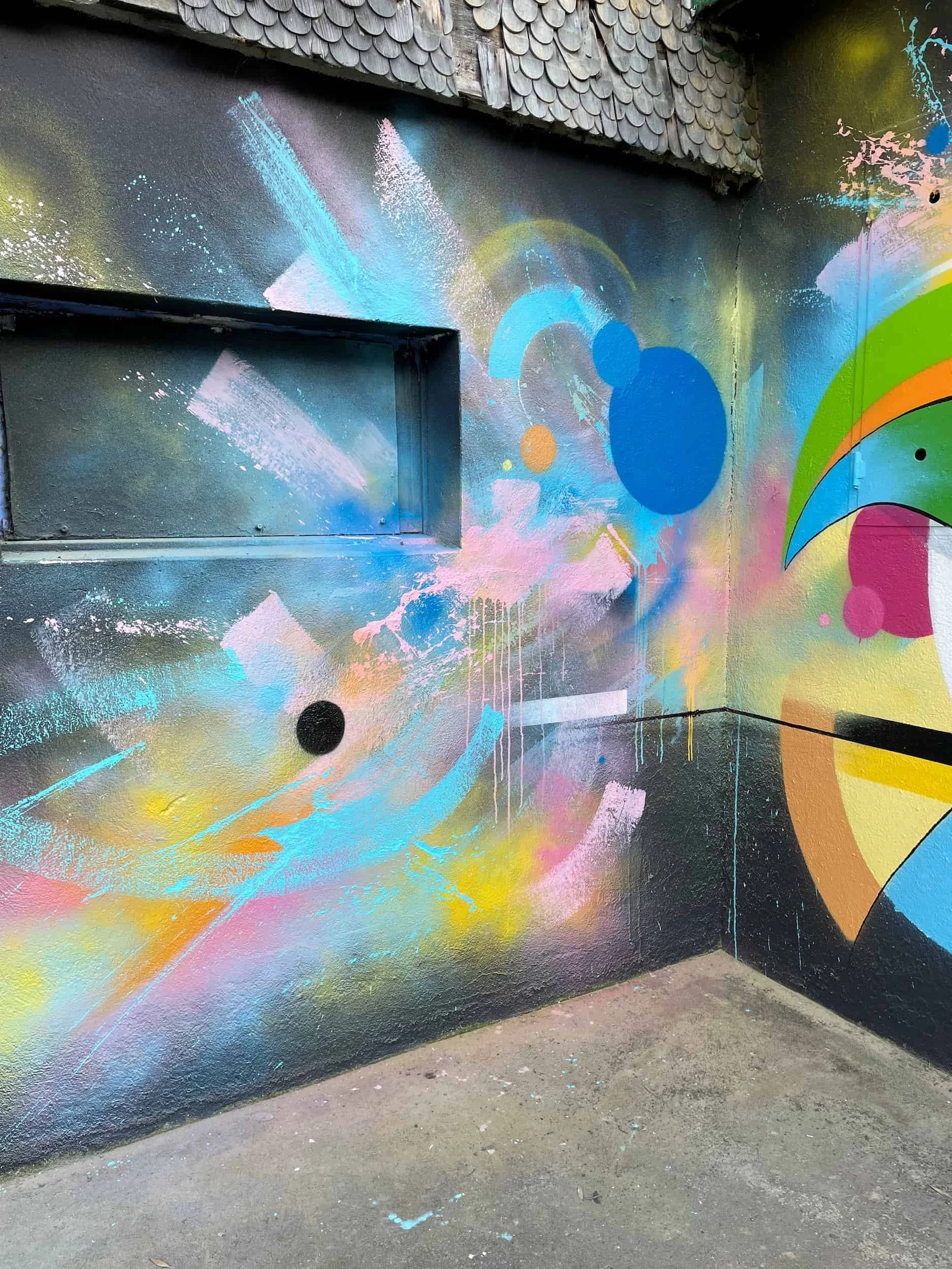 Colorful abstract graffiti mural on a black wall corner, featuring various shapes, splashes, and lines in pastel pink, yellow, green, blue, and orange.