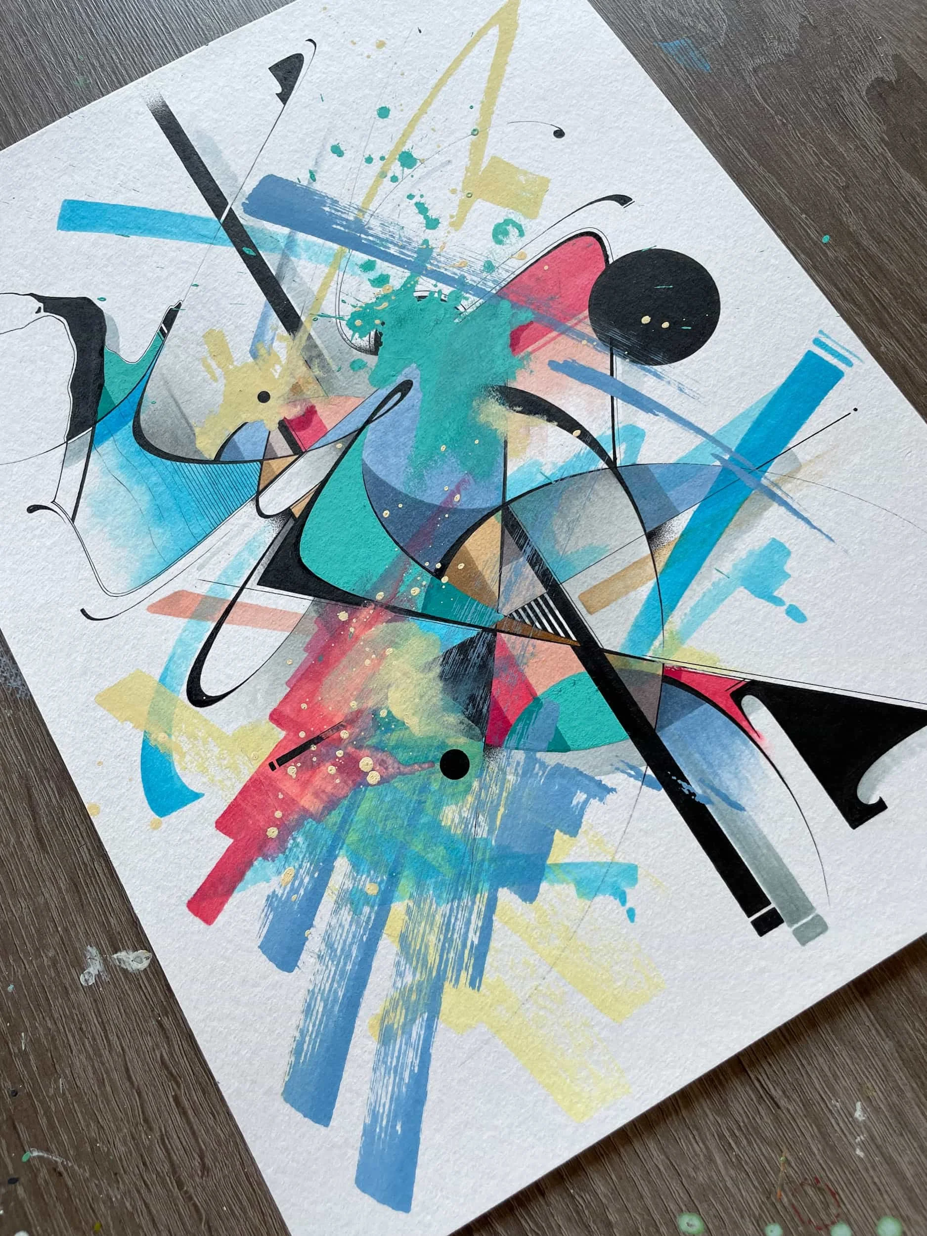 Abstract colorful painting with splashes of blue, pink, yellow, black, and gray on textured paper, placed on a wooden surface.