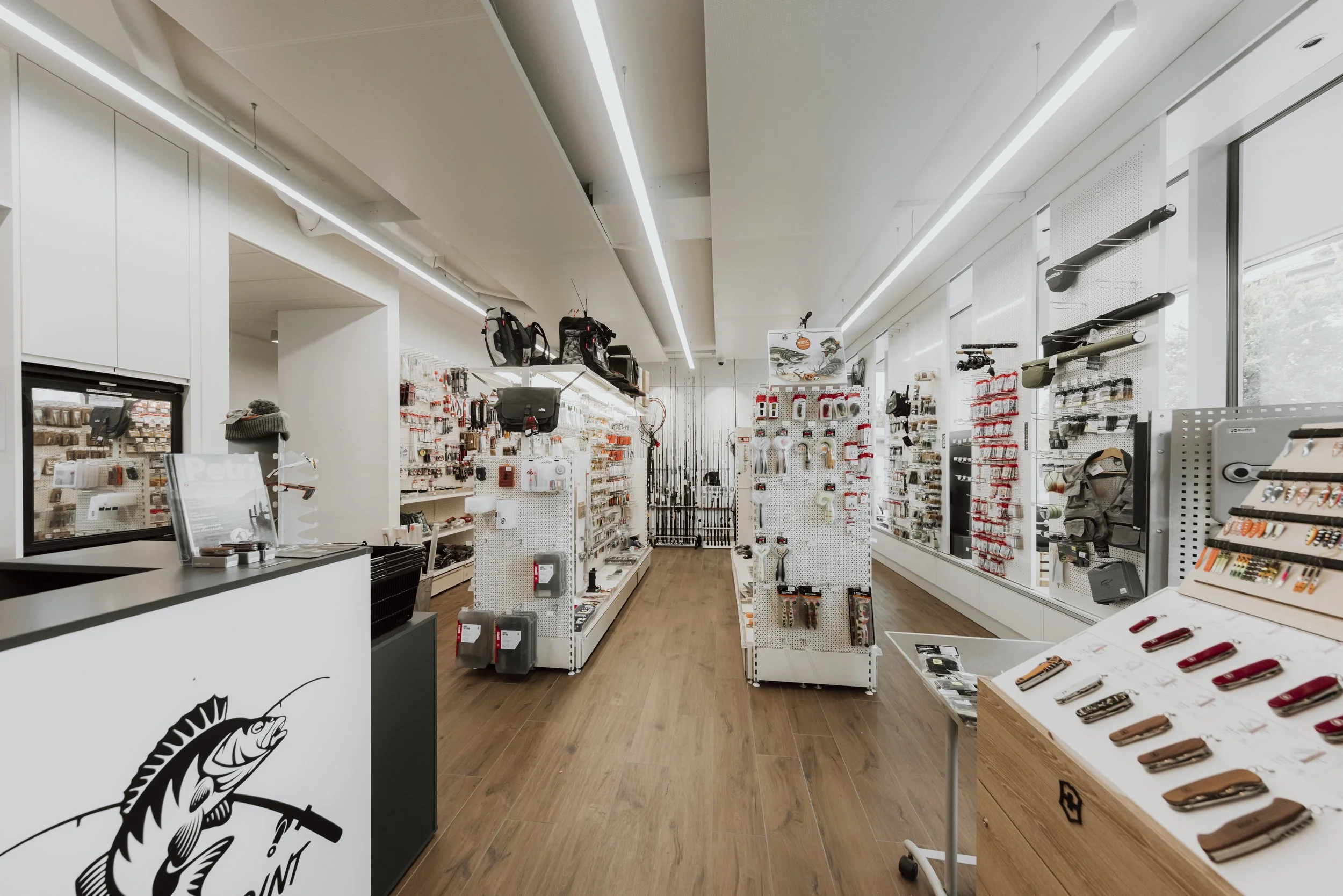 Innenansicht eines Fachgeschäfts mit Angel- und Outdoor-Produkten, weiß geflieste Wände, Holzfußboden, Regale mit Angelausrüstung, Zubehör und Messern, große Fenster an der Seite.