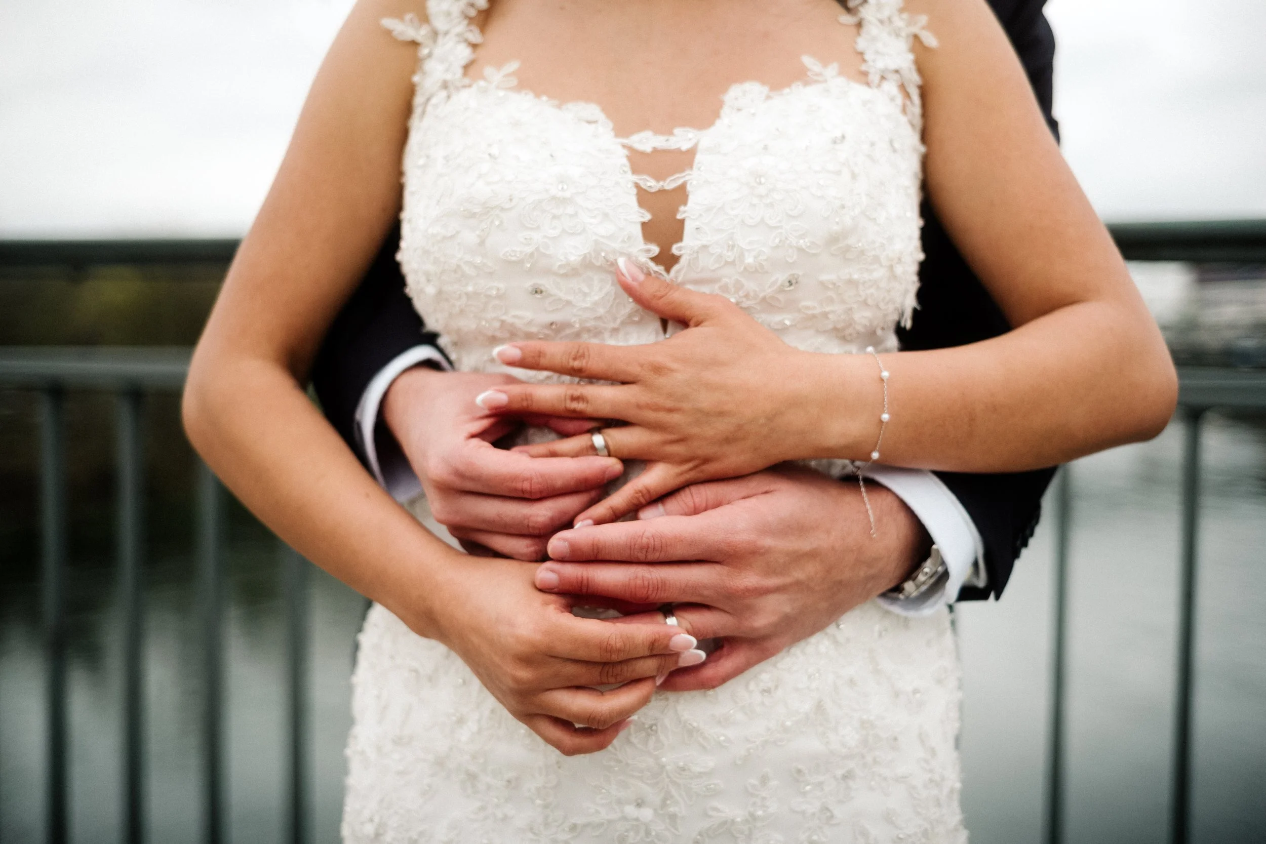 Close-up of a bride in a white lace wedding dress with floral details, embracing a groom behind her with both arms around her waist.