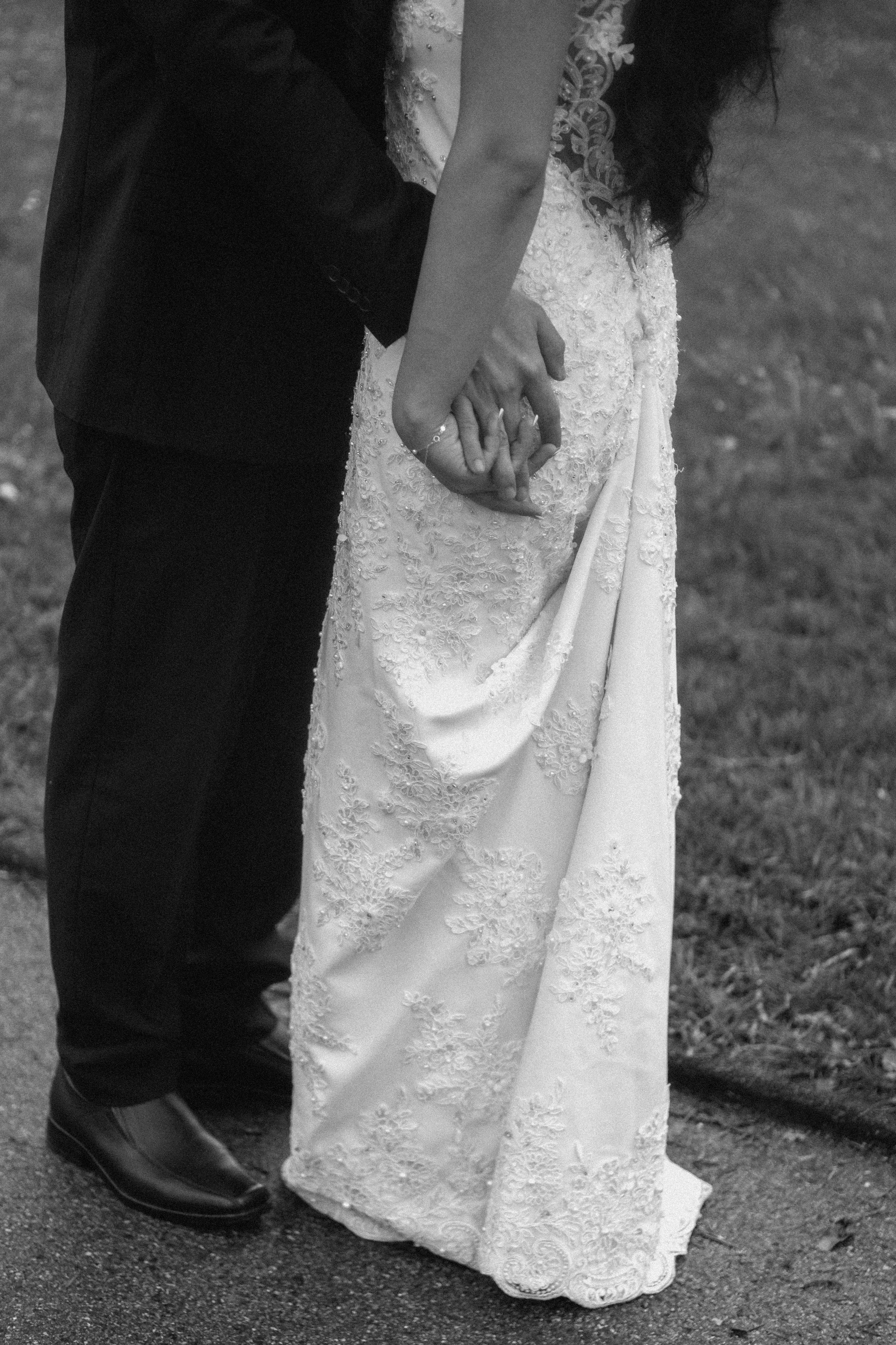 A close-up photo of a bride and groom holding hands, with only their lower bodies visible. The bride is wearing a detailed, lace wedding dress, and the groom is wearing dark trousers and polished black shoes. They are standing on a gravel path outdoo