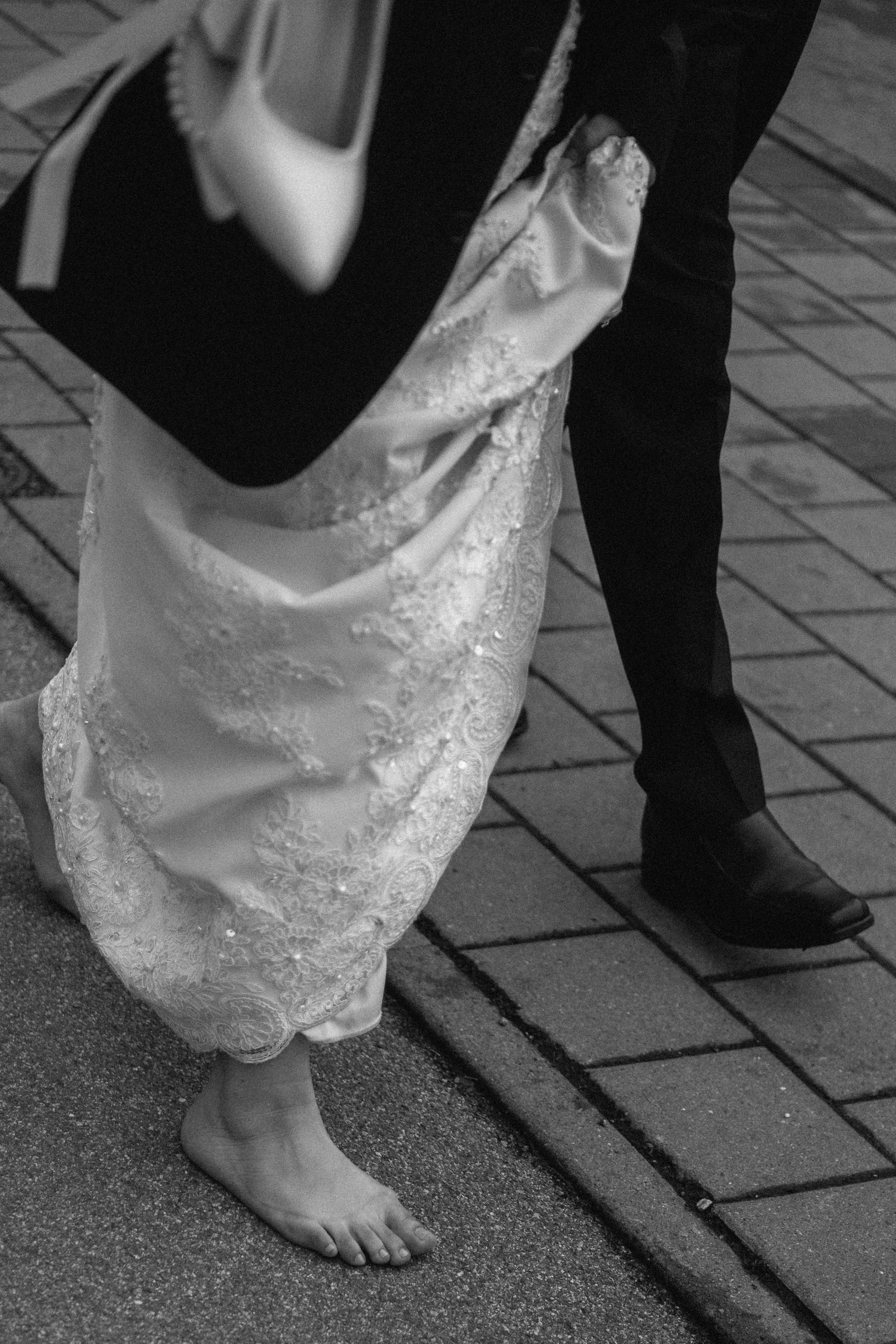 Close-up of a woman in a wedding dress standing on a sidewalk with a man in black pants and shoes visible nearby.