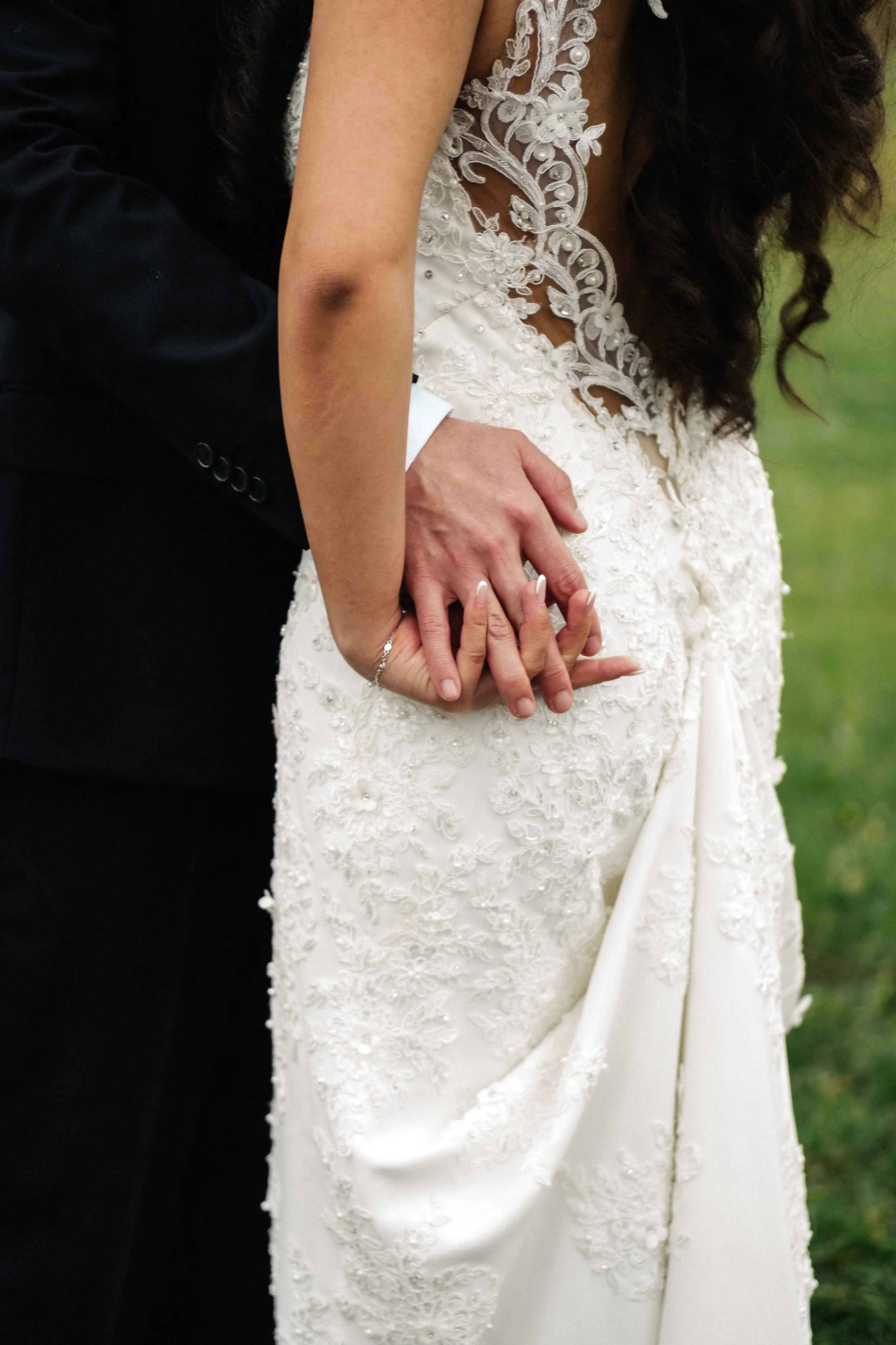 Close-up of a bride and groom holding hands, with the bride wearing an elegant lace wedding dress and the groom in a black suit, outdoors on a green background.