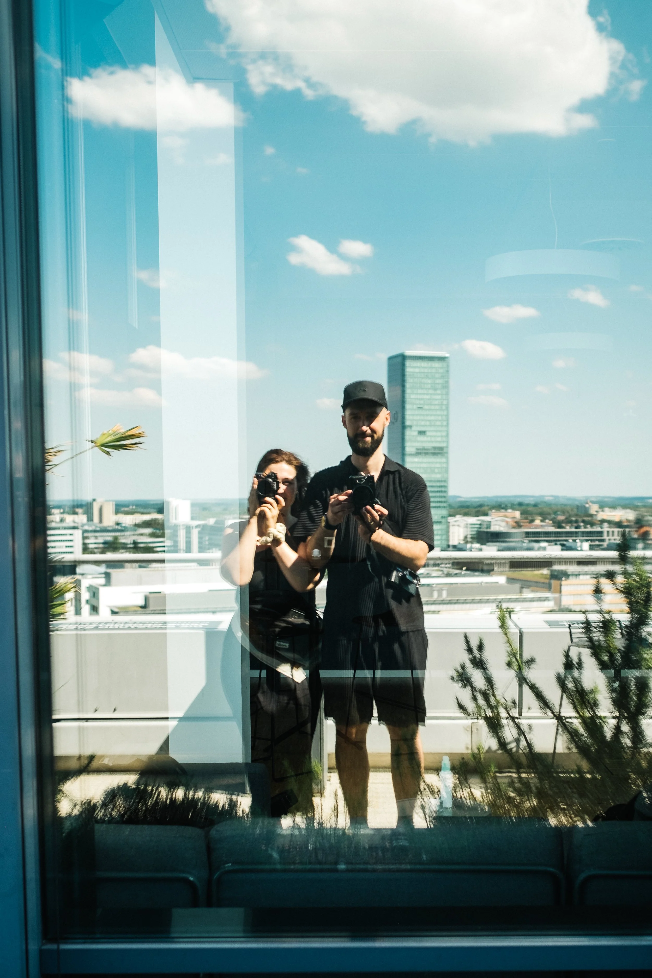 Two people taking a photo of themselves in a reflective glass window on a rooftop with a city skyline, blue sky, and some clouds in the background.