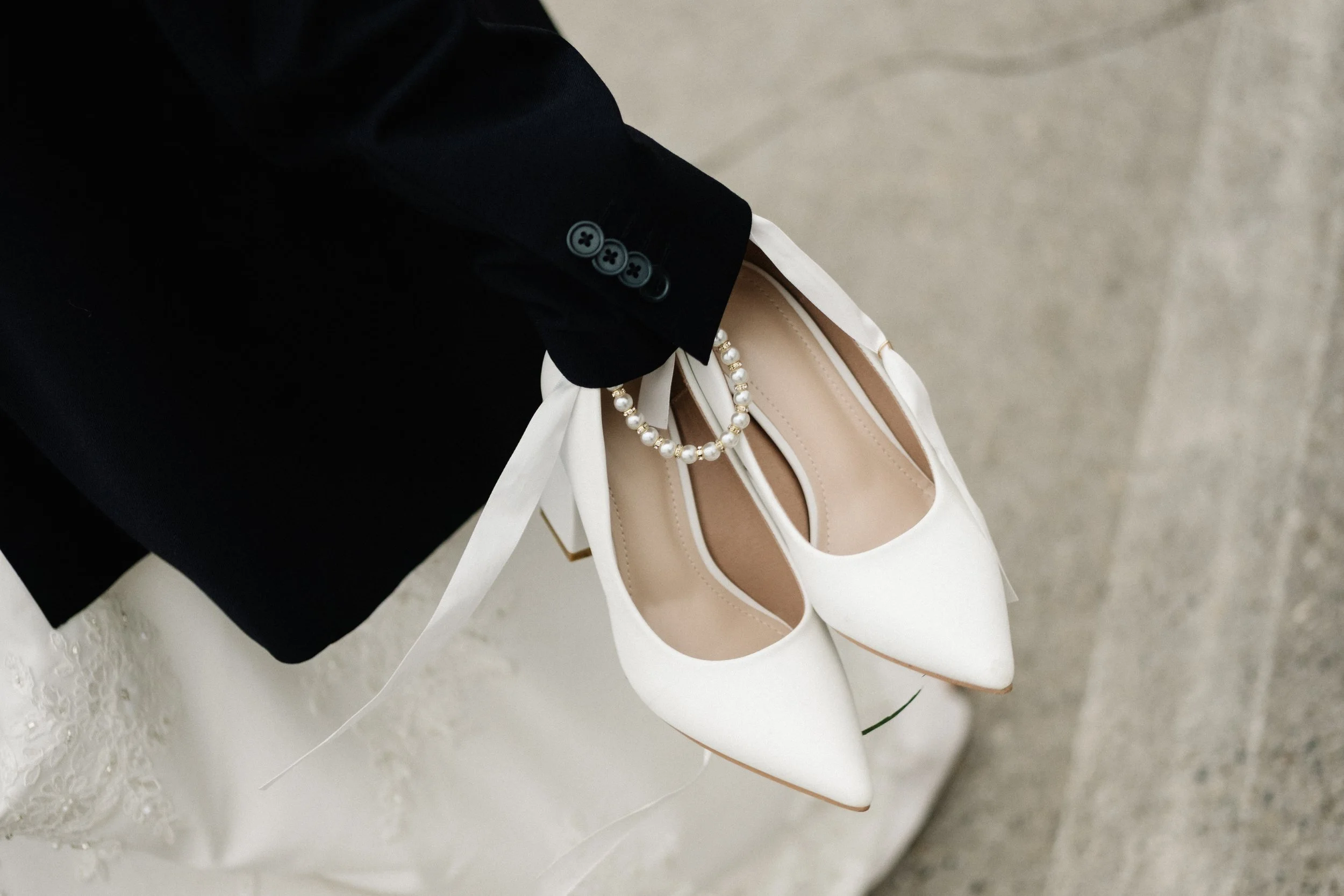 Close-up of a pair of white high heels with pearl embellishments, a white purse, and a person's hand holding the purse, with a black jacket sleeve partially visible.