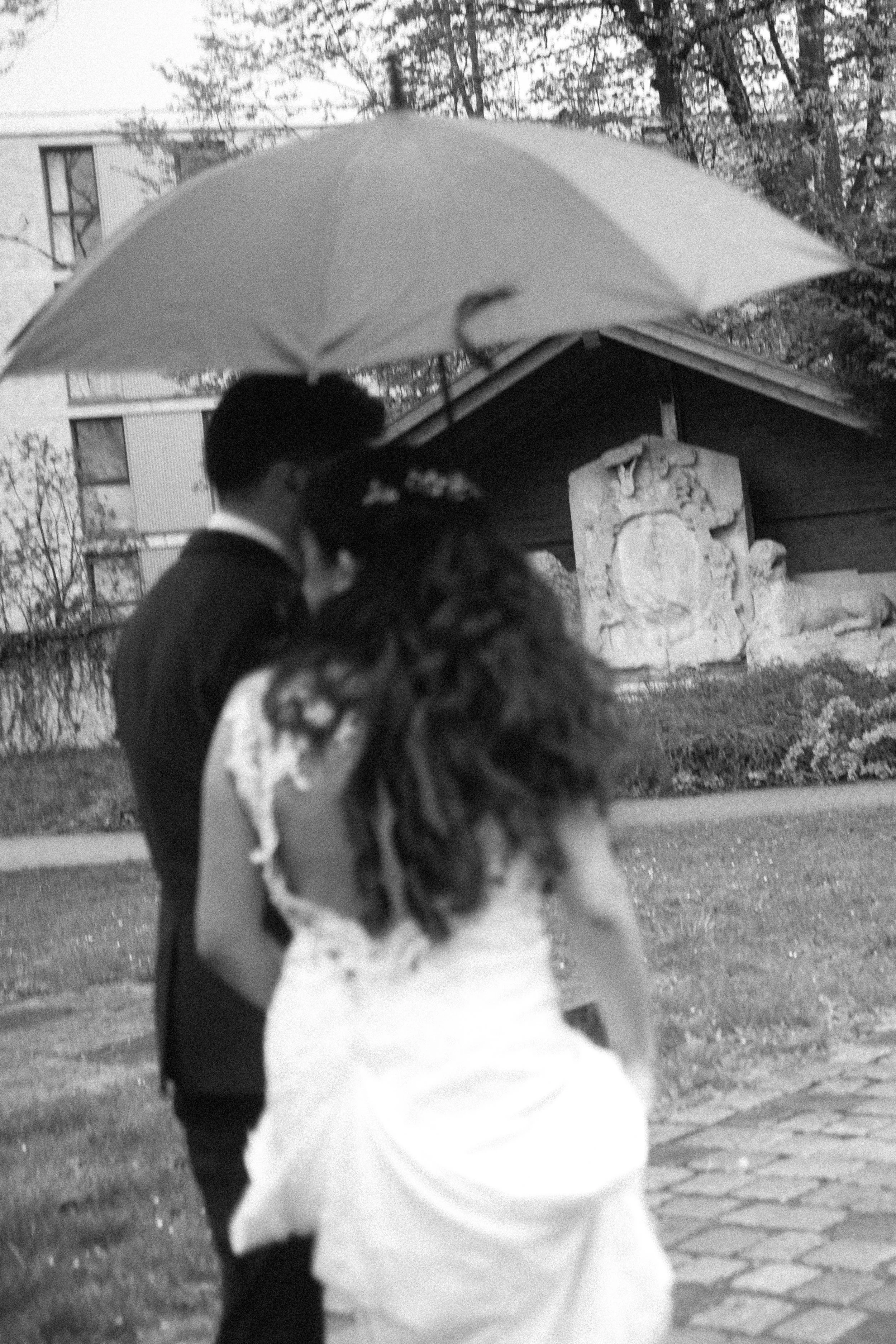 A person holding a large umbrella walking outdoors, with trees and a building in the background.