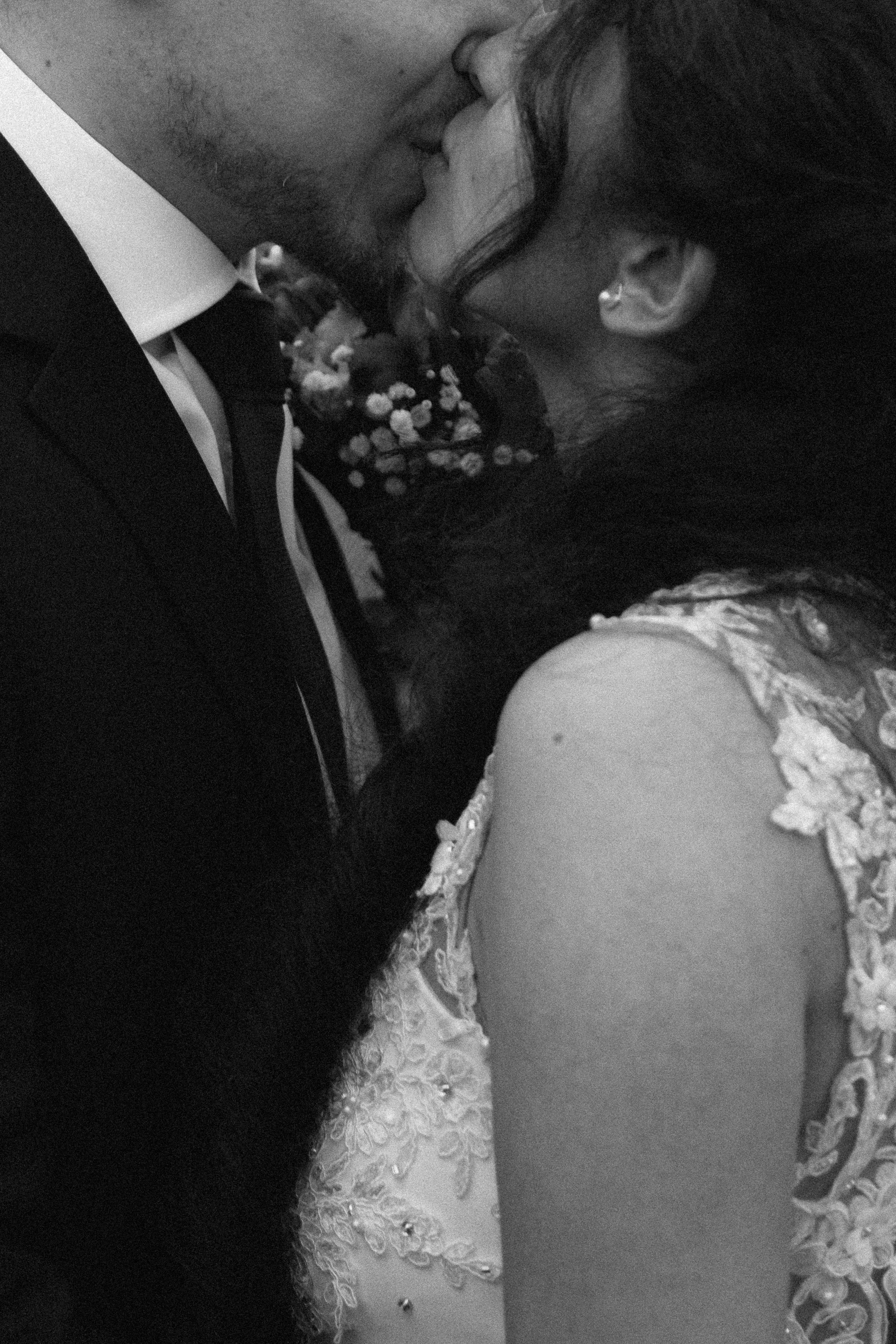 Close-up of a couple kissing, with the woman wearing a wedding dress with lace details and the man dressed in a black tuxedo with a white shirt and black bow tie.