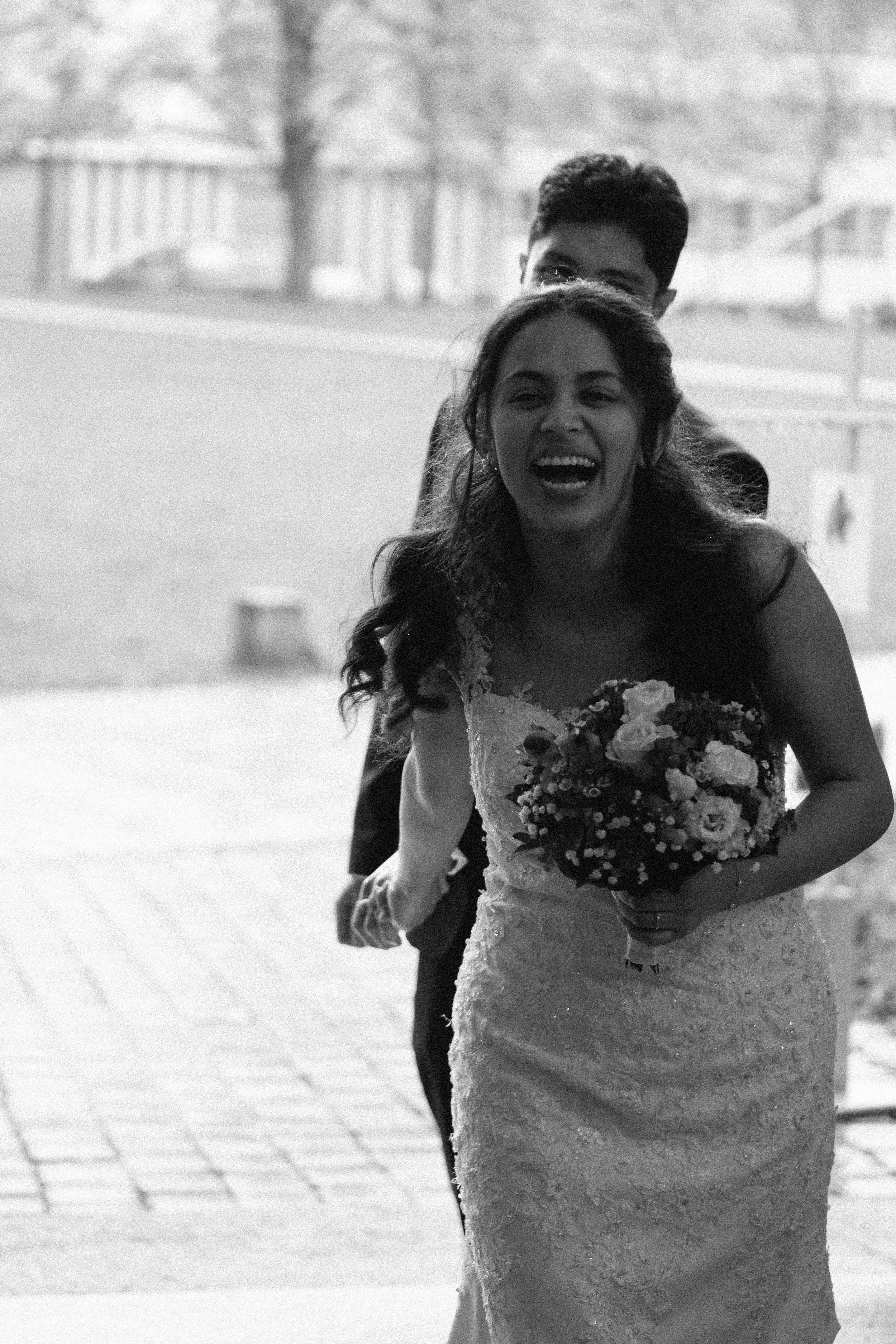 A bride and groom are walking outside, with the bride holding a bouquet of flowers and smiling. The groom is partially visible behind her, and both are dressed in wedding attire.