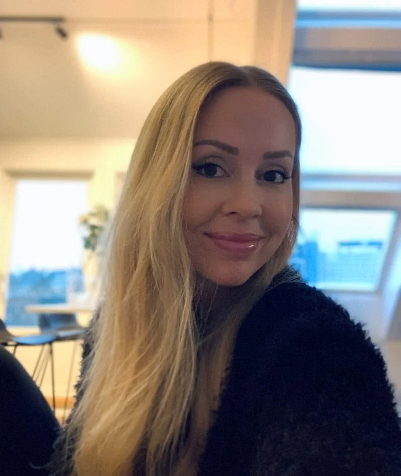 Kvinne med langt blondt hår, i et innendørsmiljø med vinduer i bakgrunnen.