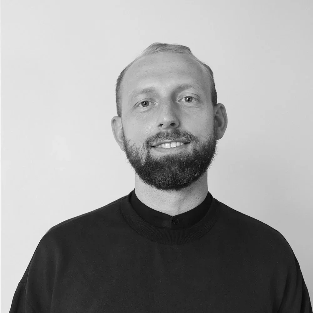 Schwarz-weißPorträt eines Mannes mit Bart, short Haar und dunklem Pullover, lächelnd, vor hellem Hintergrund.