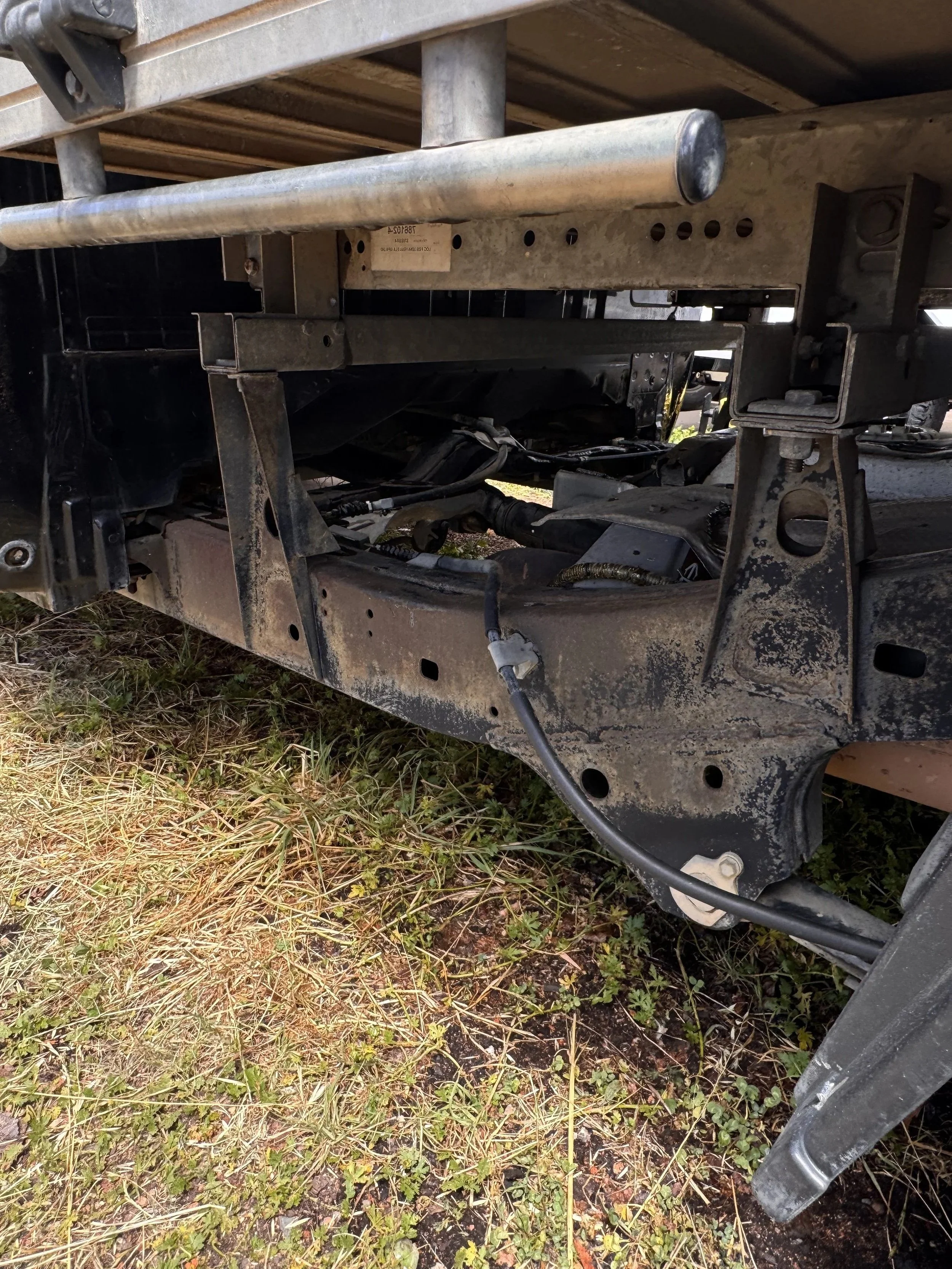 Close-up of the underside of a vehicle showing its rusty metal frame and chassis components, with some dirt and grass on the ground underneath.