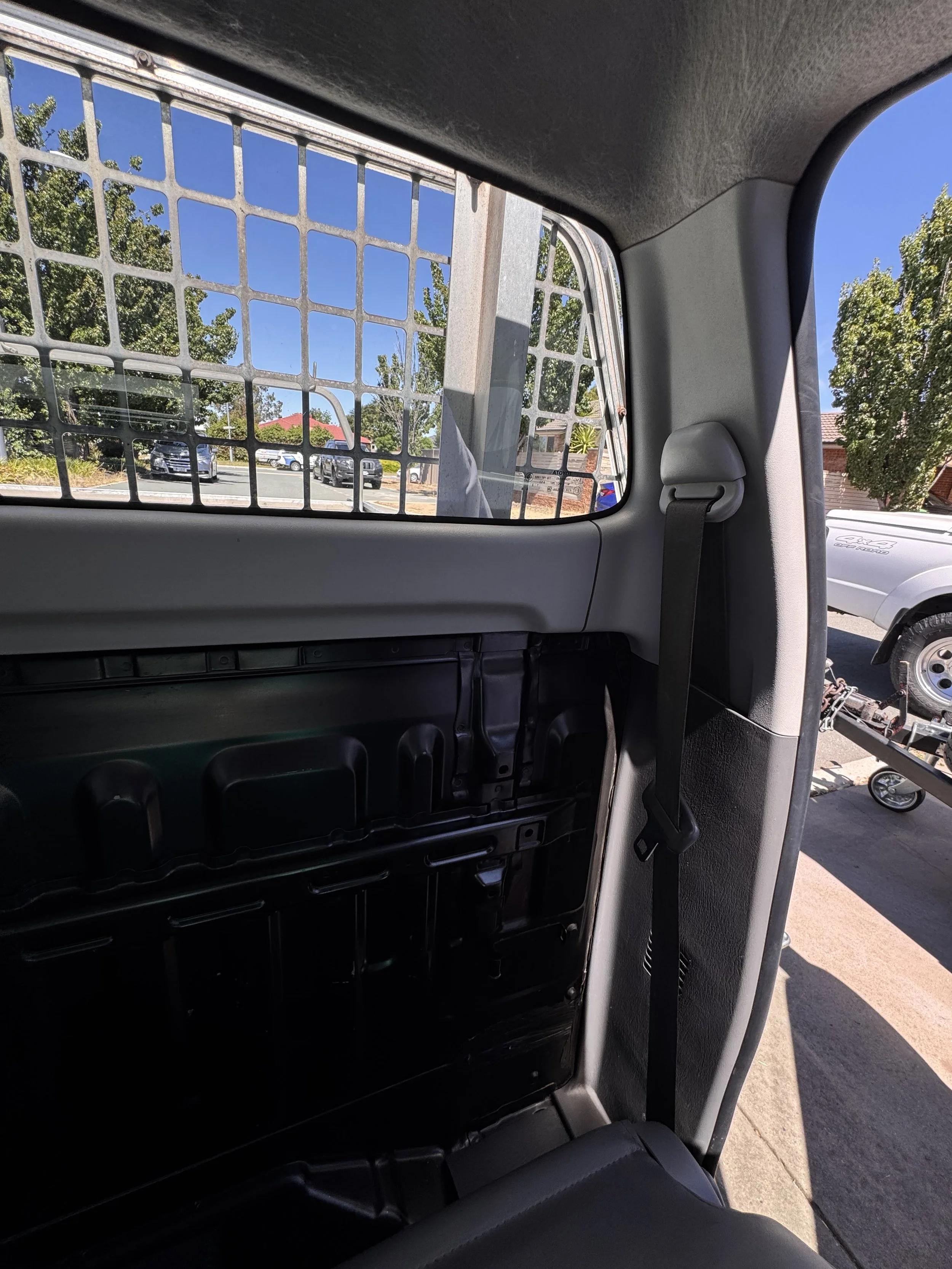 Inside the back of a utility vehicle with a metal grid partition, seatbelt, and a view of parked cars, trees, and a sidewalk outside.