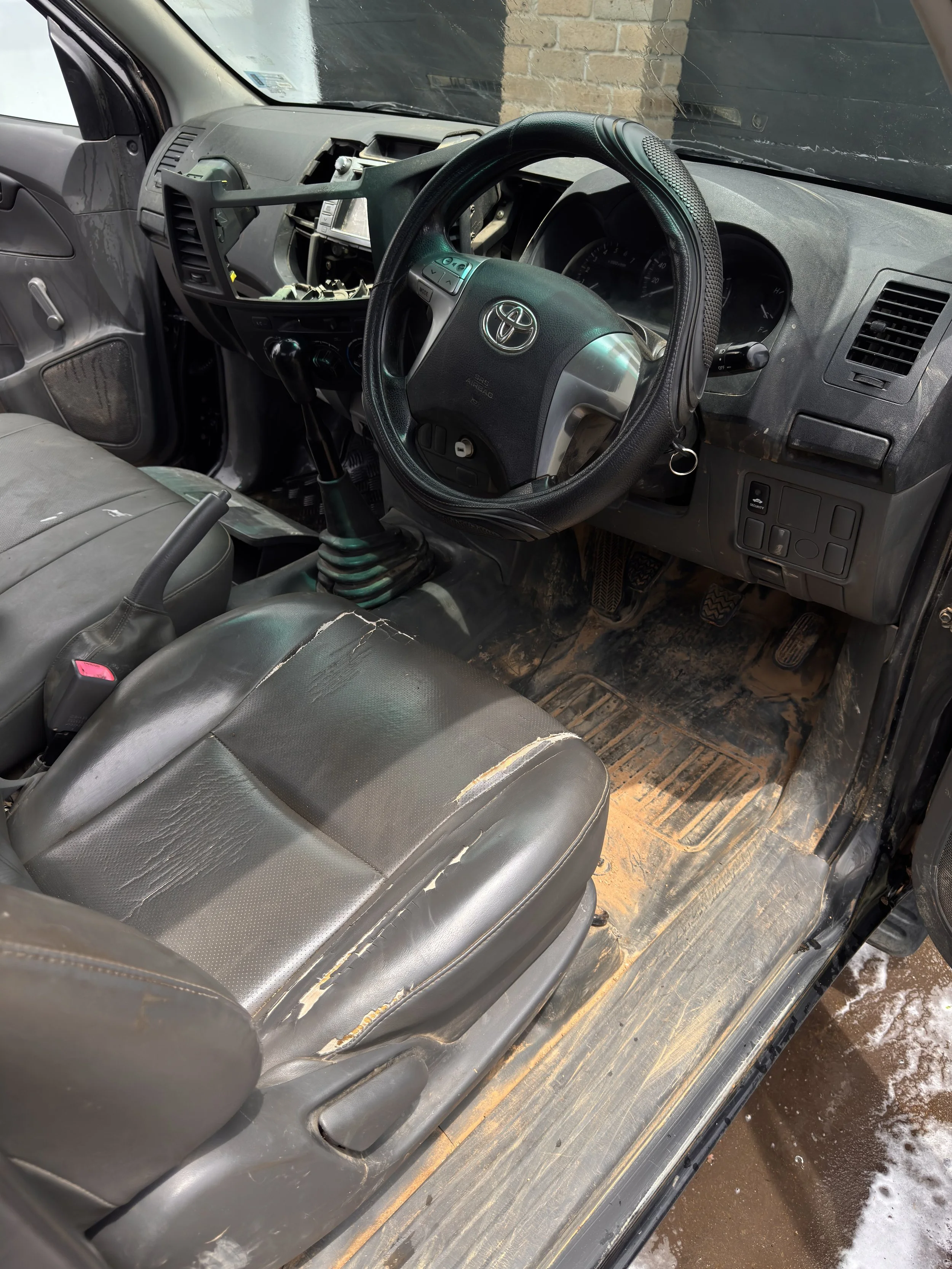 The interior of a dirty and damaged vehicle, showing the driver's seat with torn upholstery, a steering wheel, and a dashboard with some missing components.
