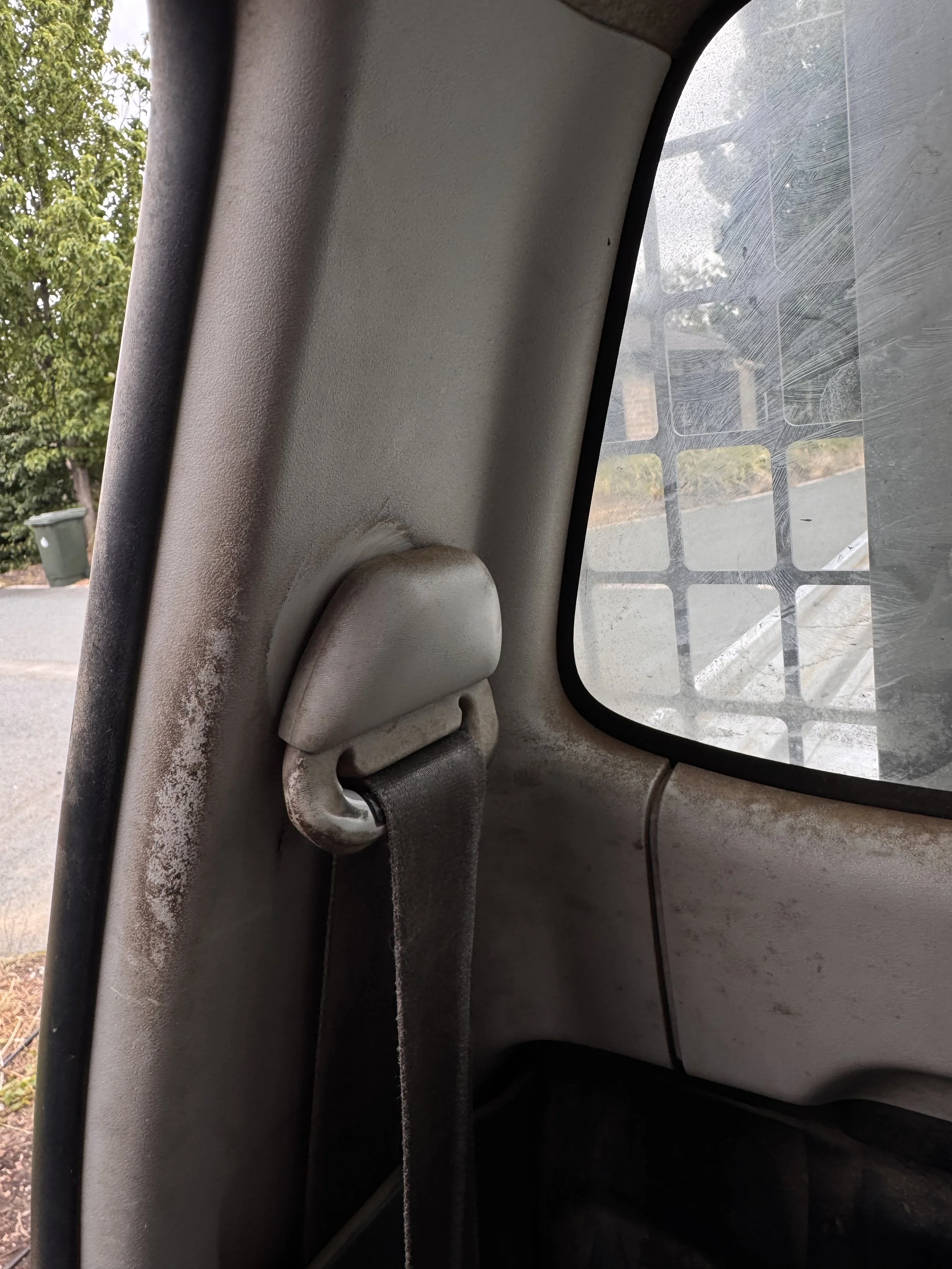 The interior of a vehicle showing a grey seatbelt anchored to the side, with a cloudy window and outdoor scenery including trees, a street, and a green trash bin.