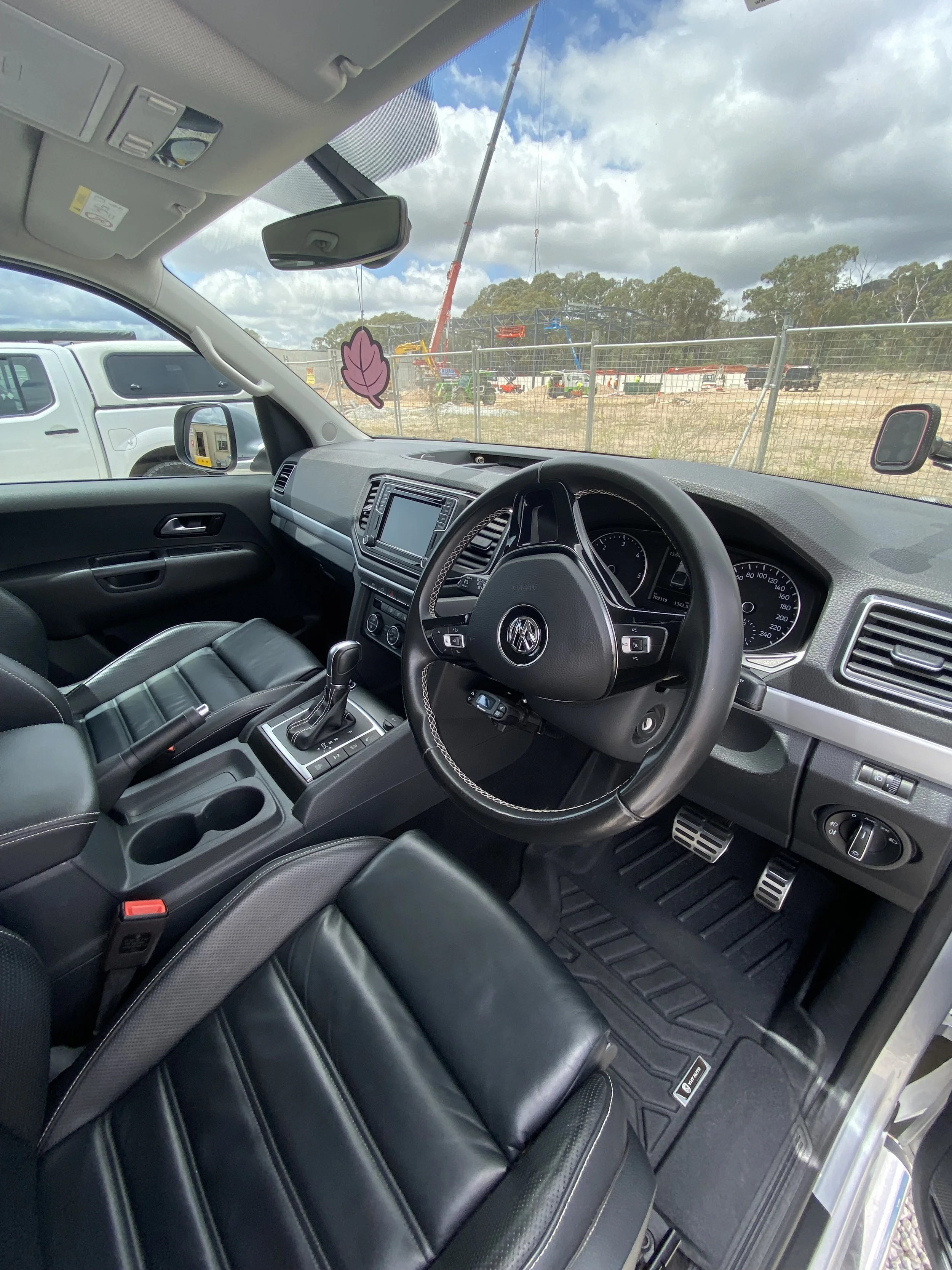 Interior view of a Volkswagen vehicle parked at a construction site, showing the steering wheel, dashboard, gear shift, black leather seats, and a pink leaf air freshener hanging from the rearview mirror, with construction equipment and fencing visib