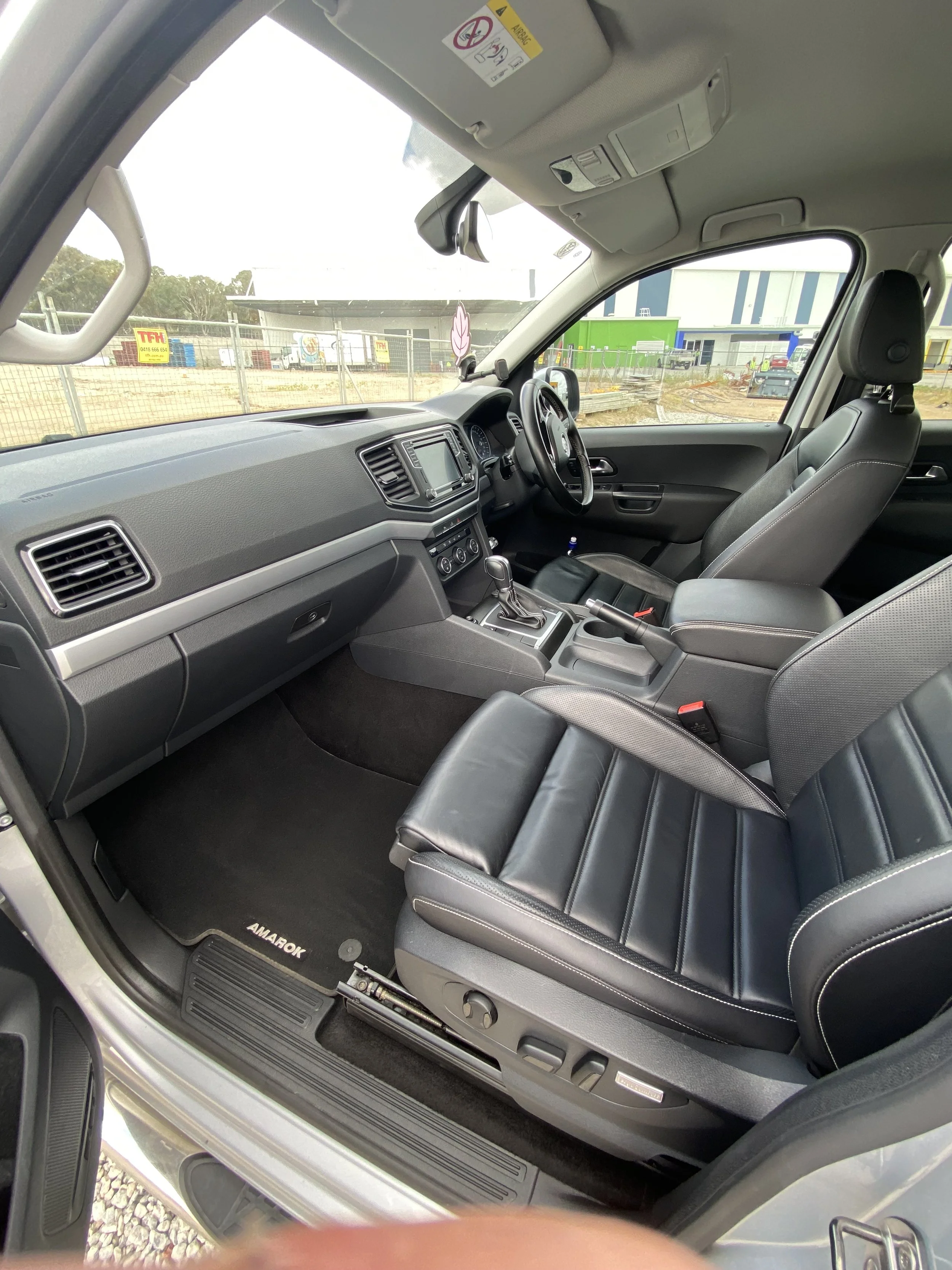 Interior view of a vehicle showing the driver and front passenger seat, dashboard, steering wheel, and gear shift, with a construction site visible through the windshield.