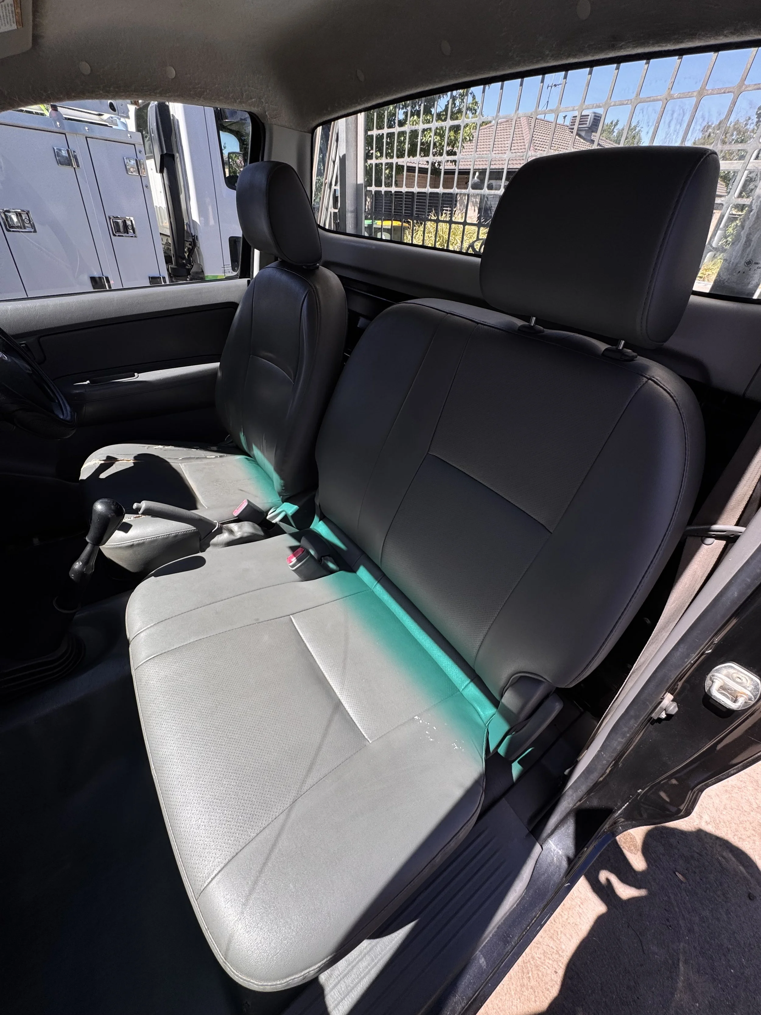 The interior of a vehicle showing the front and back seats with black upholstery, a gear shift, and a window with a barred metal grid outside.
