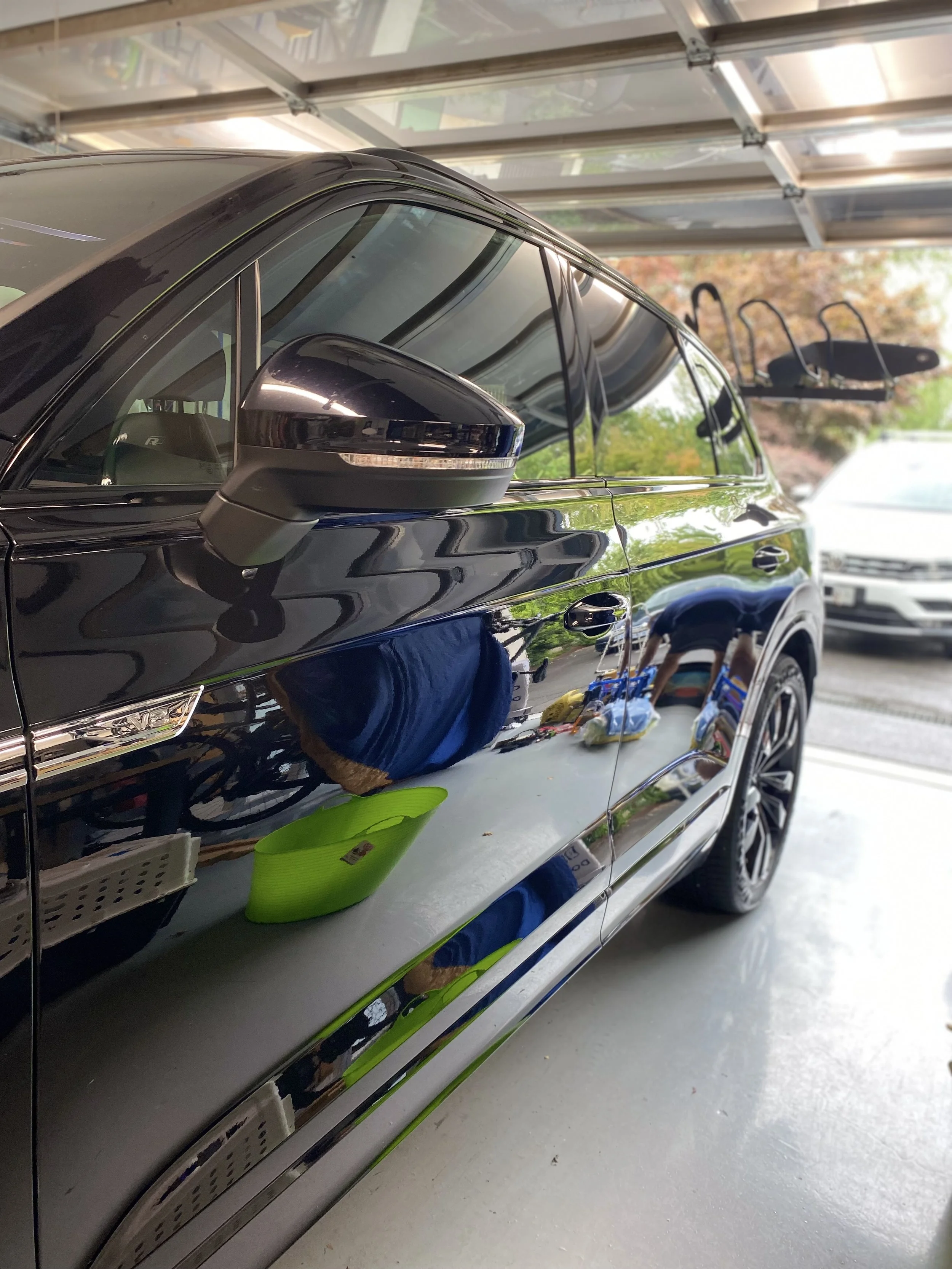 A shiny black car parked inside a garage, with reflections showing a person in blue clothes, a green basket, and various garage items including a bicycle and a white vehicle outside.