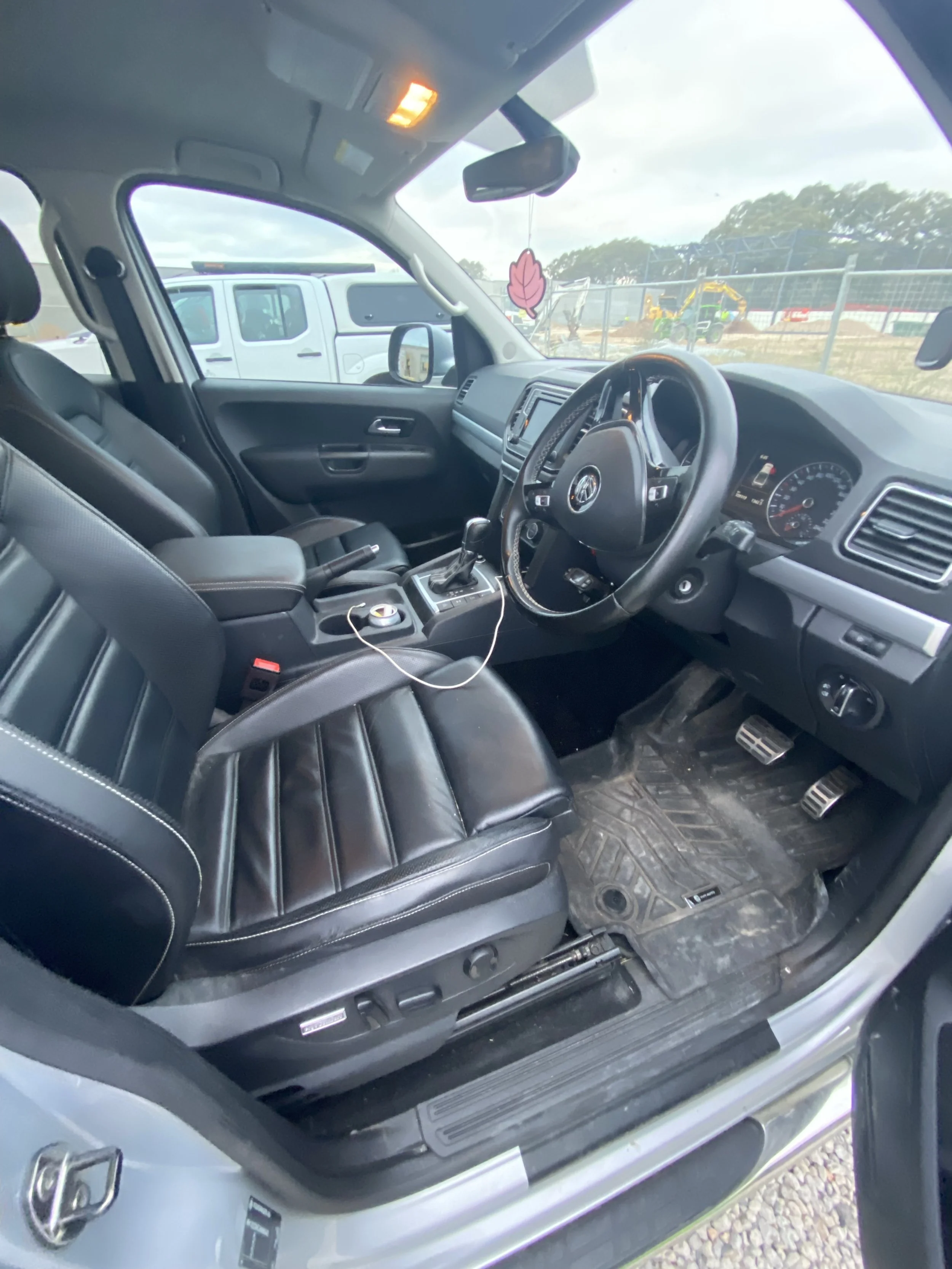 Interior of a vehicle showing black leather front seats, a dashboard with a steering wheel, gear shifter, and various controls. The driver's side floor has a dirtied rubber mat and pedals are visible. Outside, other vehicles and construction equipmen