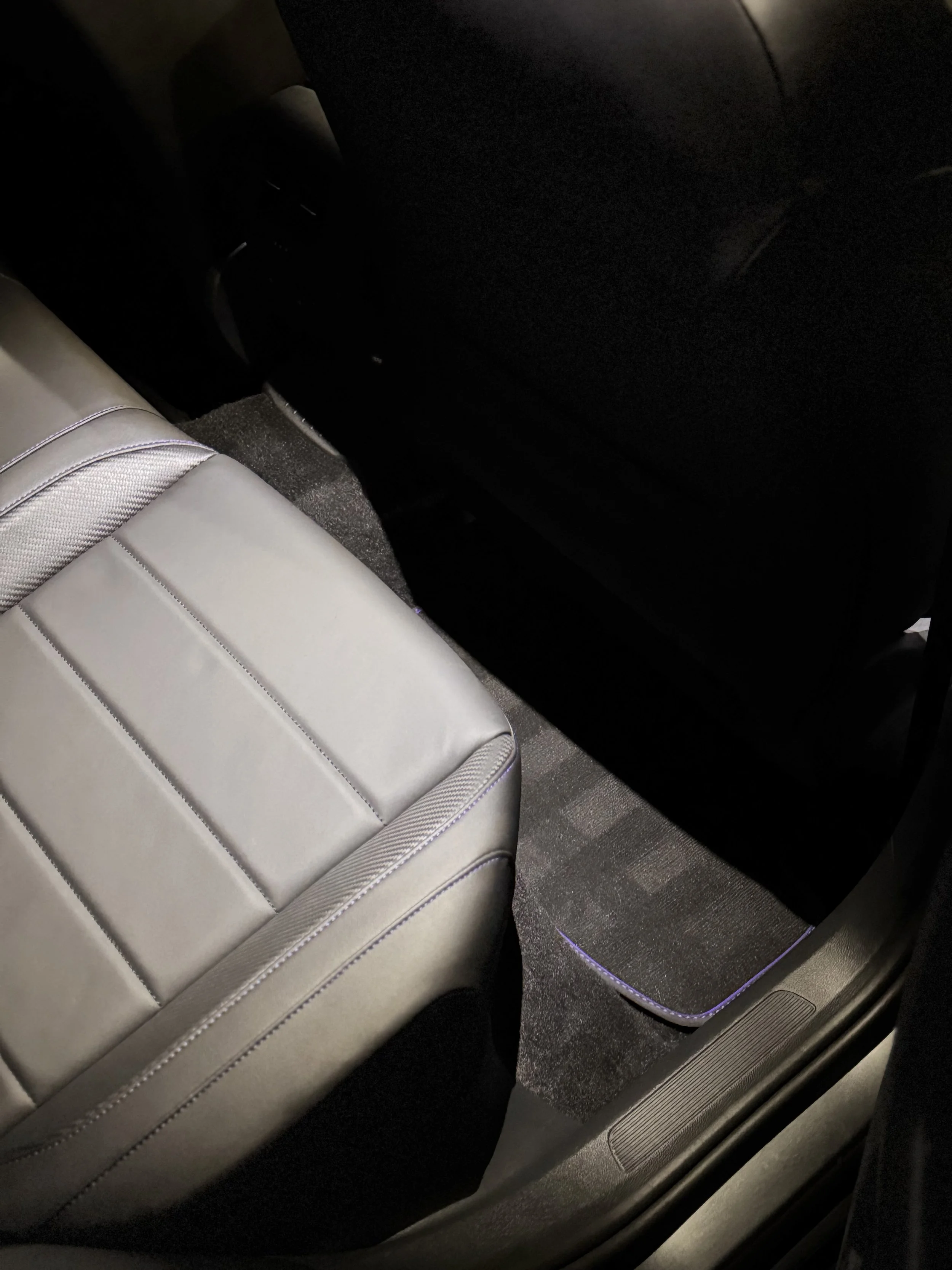 The backseat of a vehicle showing a blackleather seat, black carpeting, and part of the back of a black front seat.