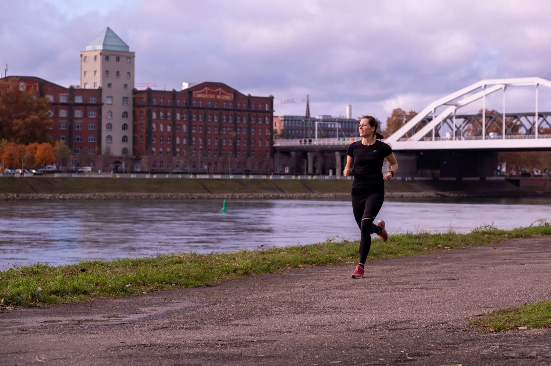 Eine Frau läuft joggend am Flussufer entlang bei bewölktem Himmel, im Hintergrund sind Stadtgebäude und eine Brücke sichtbar.