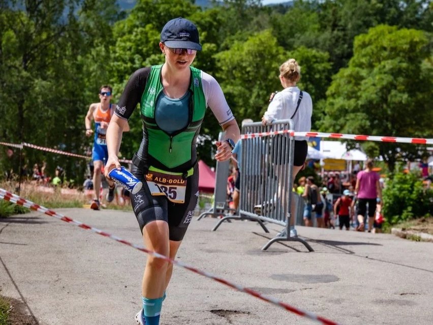 Eine Frau läuft bei einem Triathlon, trägt eine grüne und schwarze Wettkampfkleidung, eine Mütze, Sonnenbrille und hat eine Wasserflasche in der Hand, im Hintergrund sind andere Teilnehmer und Zuschauer.