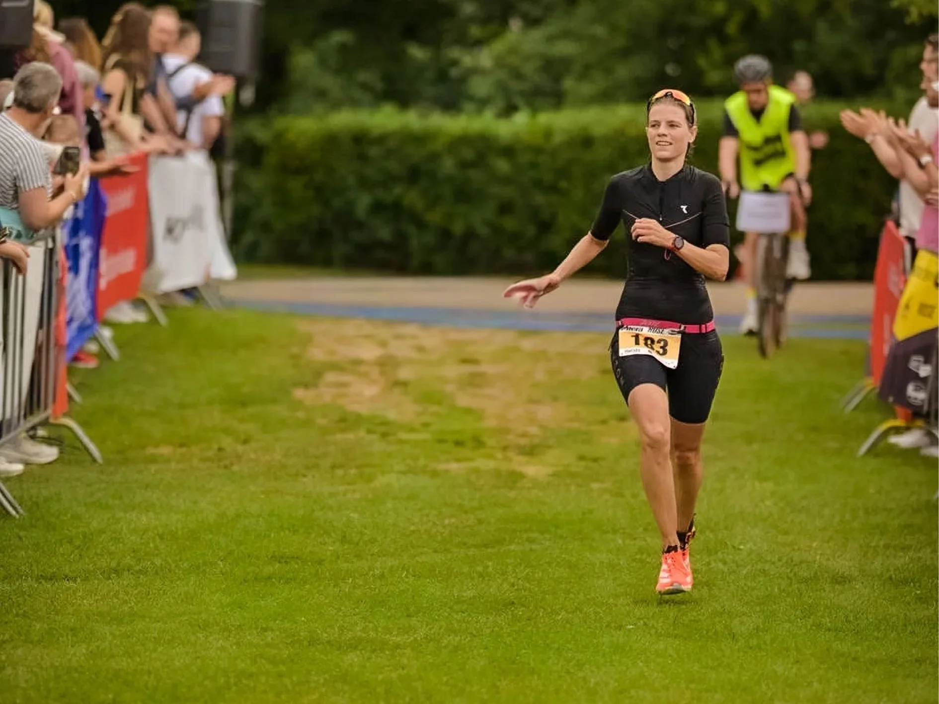 Frau beim Sieg beim Laufwettbewerb, Läufer mit Startnummer 183, jubelnde Zuschauer am Rand, im Freien, grüne Bäume im Hintergrund.