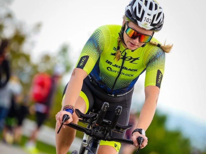Frau in Fahrradkleidung beim Radfahren, trägt Helm und Sonnenbrille, neben ihr sind andere Radfahrer im Hintergrund.
