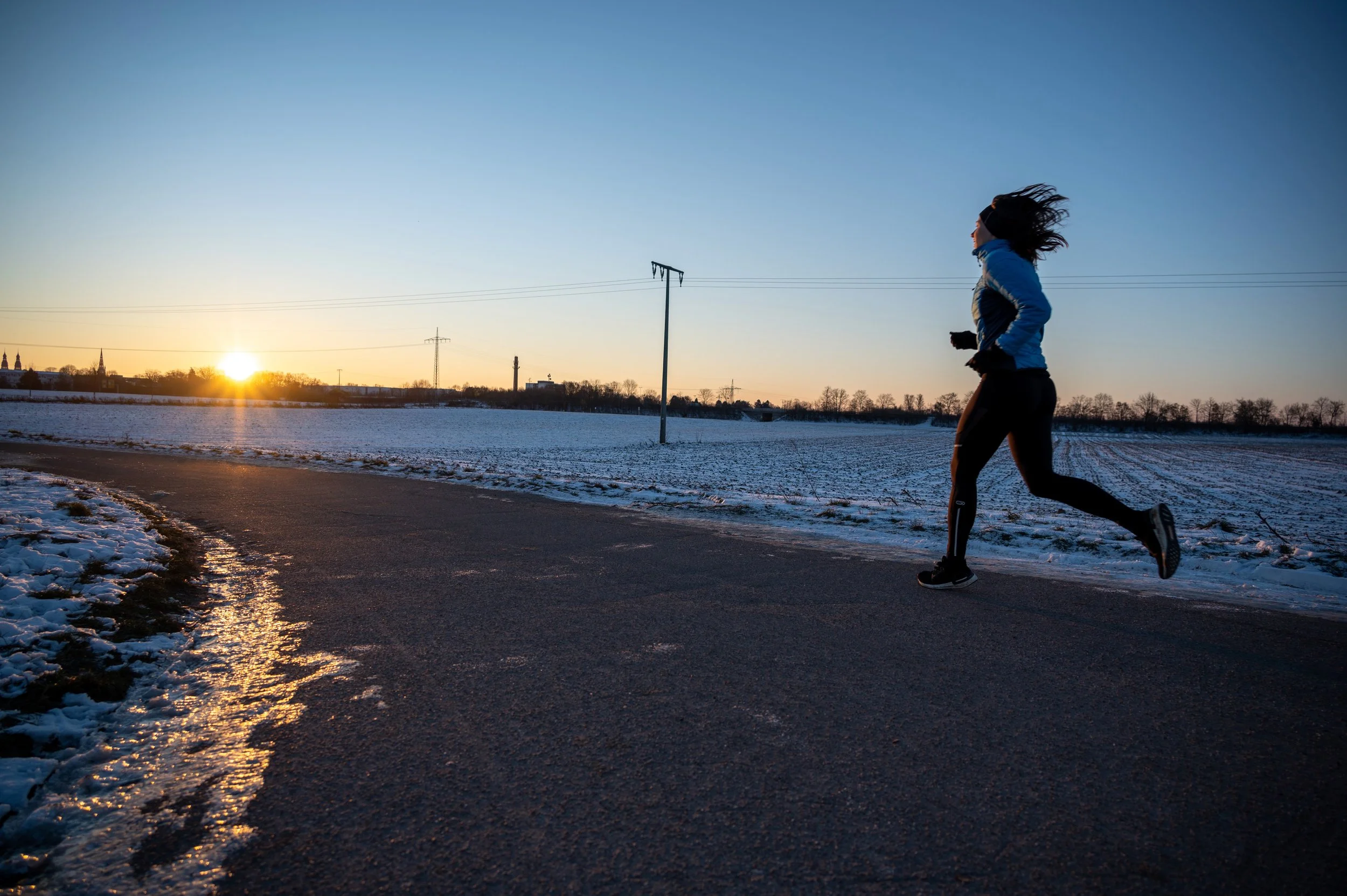 Eine Frau joggt bei Sonnenuntergang auf einer asphaltierten Straße im Winter, mit Schnee am Rand, in einer ländlichen Gegend mit Feldern und Stromleitungen im Hintergrund.