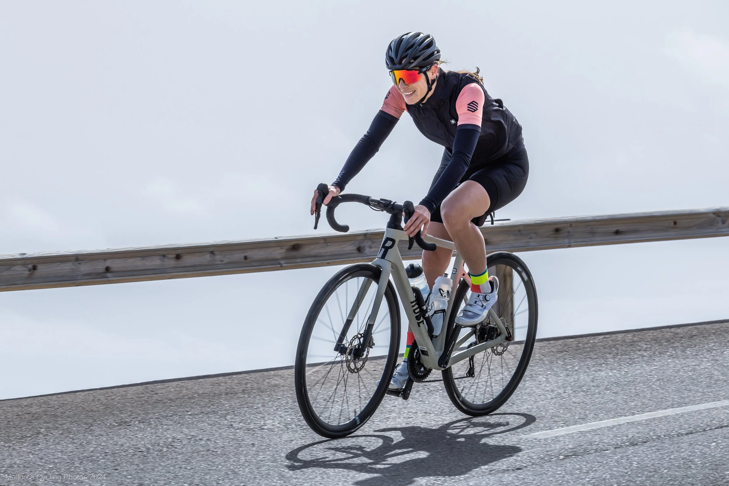 Frau fährt mit Rennrad bergauf auf einer Straße mit Holzschutzplanke bei bewölktem Himmel.
