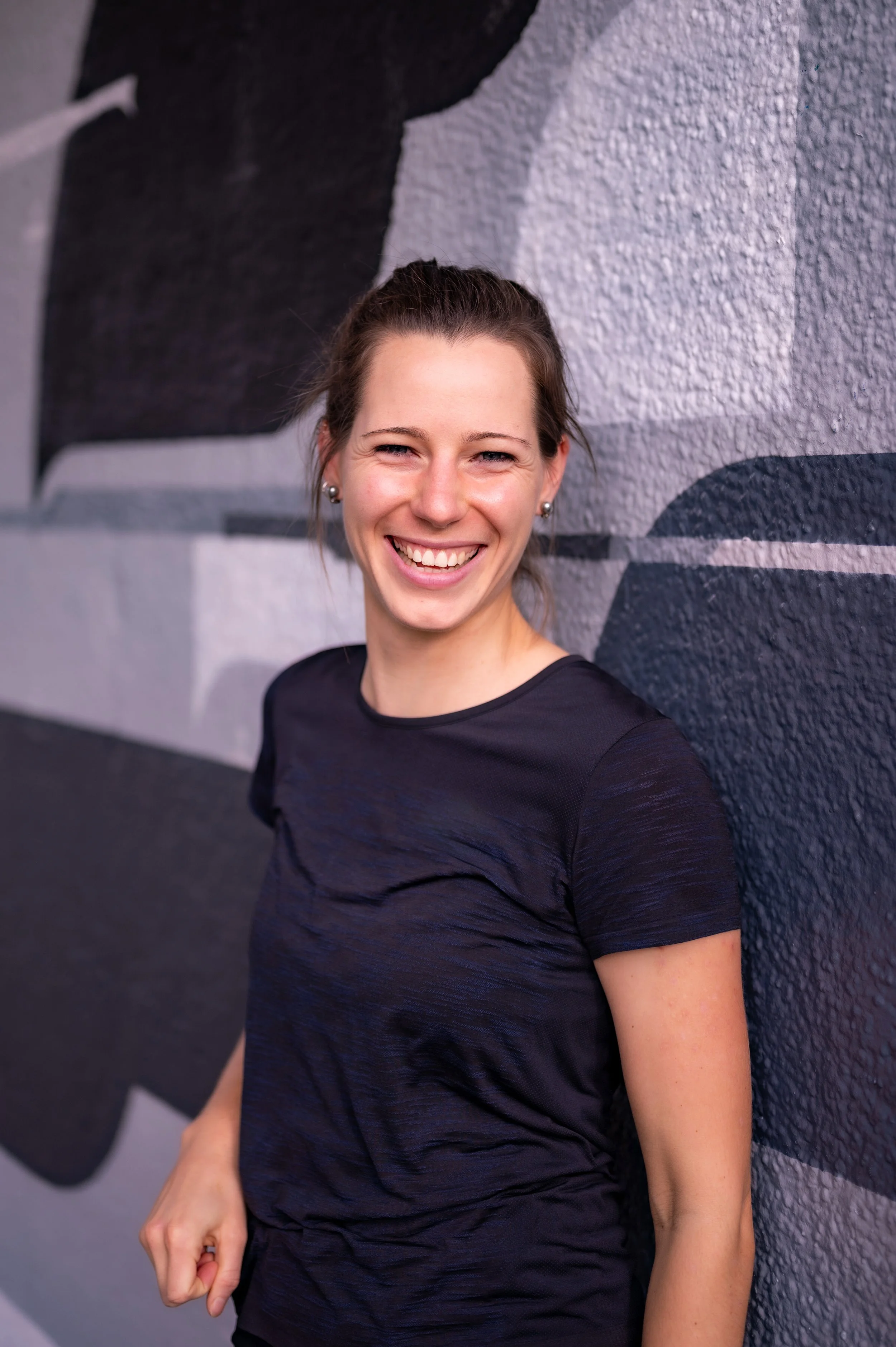 Eine lächelnde junge Frau mit braunen Haaren, die einen schwarzen Sport-Trainingsshirt trägt, posiert vor einer Graffiti-Wand.
