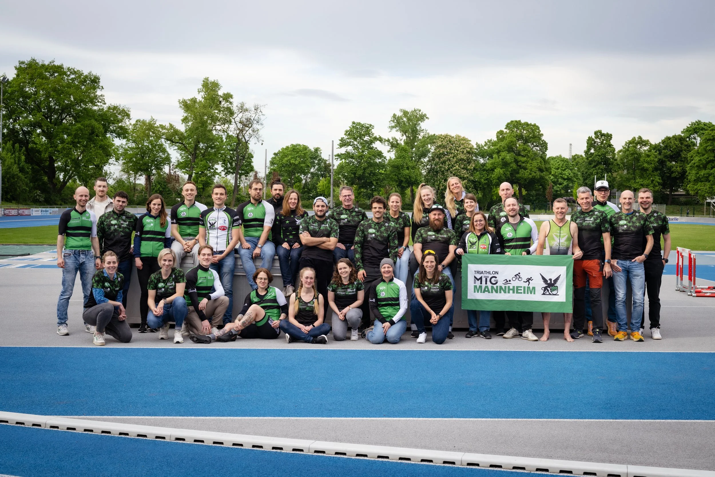 Gruppe von Menschen beim Triathlon-Event in Mannheim, einige in grünen und schwarzen Sportoutfits, einige halten ein Banner mit dem Text "Triathlon MTG Mannheim". Sie stehen auf einer Laufbahn mit blauen Bahnen, im Hintergrund sind Bäume und eine Laufbahn zu sehen.