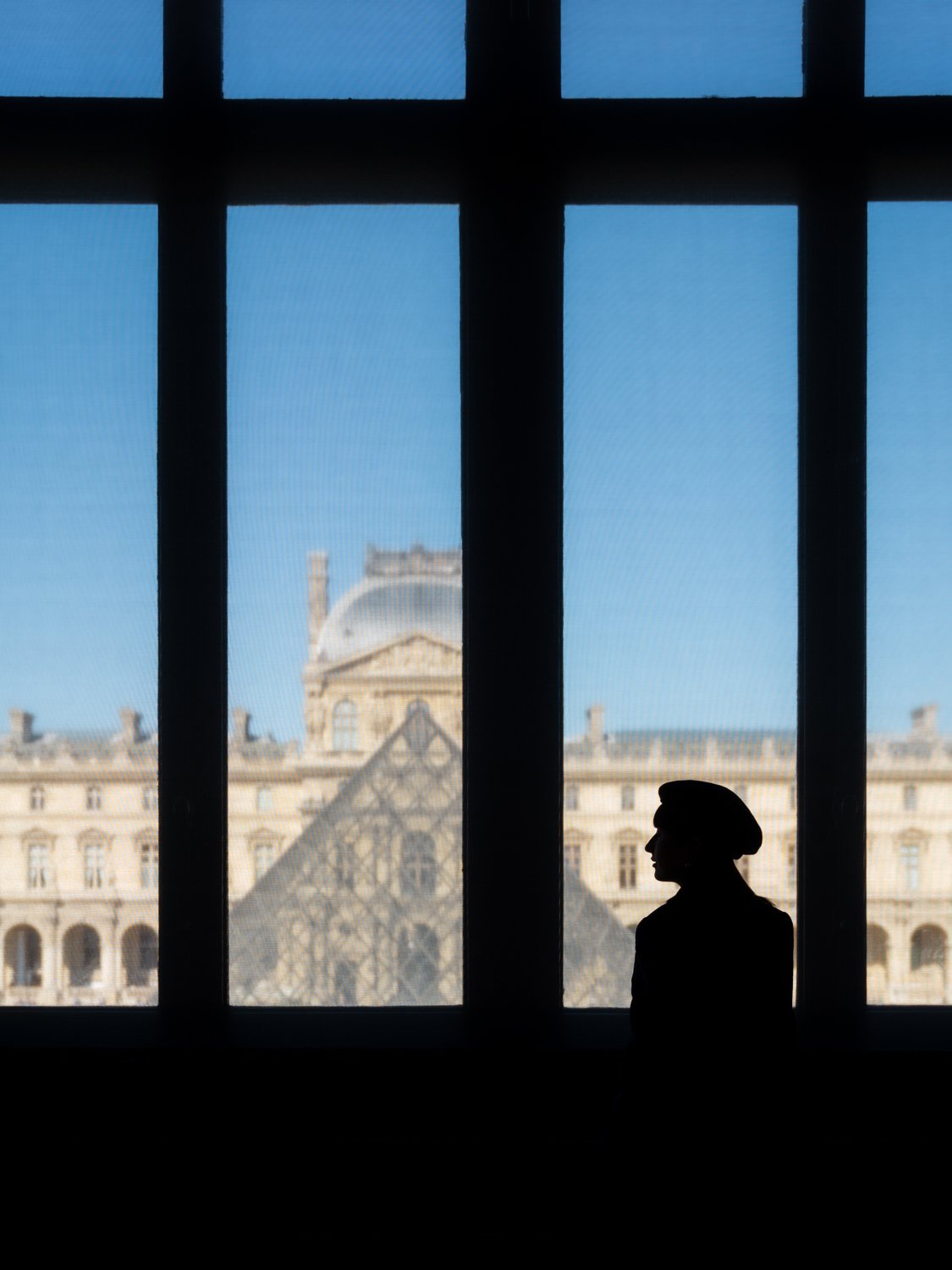 Silhouette of a woman wearing a hat, looking out of a large window with view of the Louvre Museum in Paris, France, during daytime.