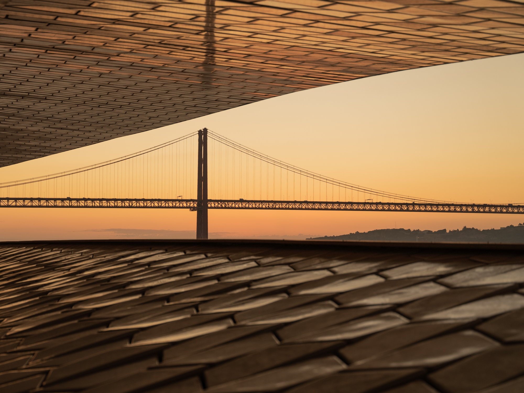 A bridge seen through framed openings of a building at sunset, with a tiled roof in the foreground.
