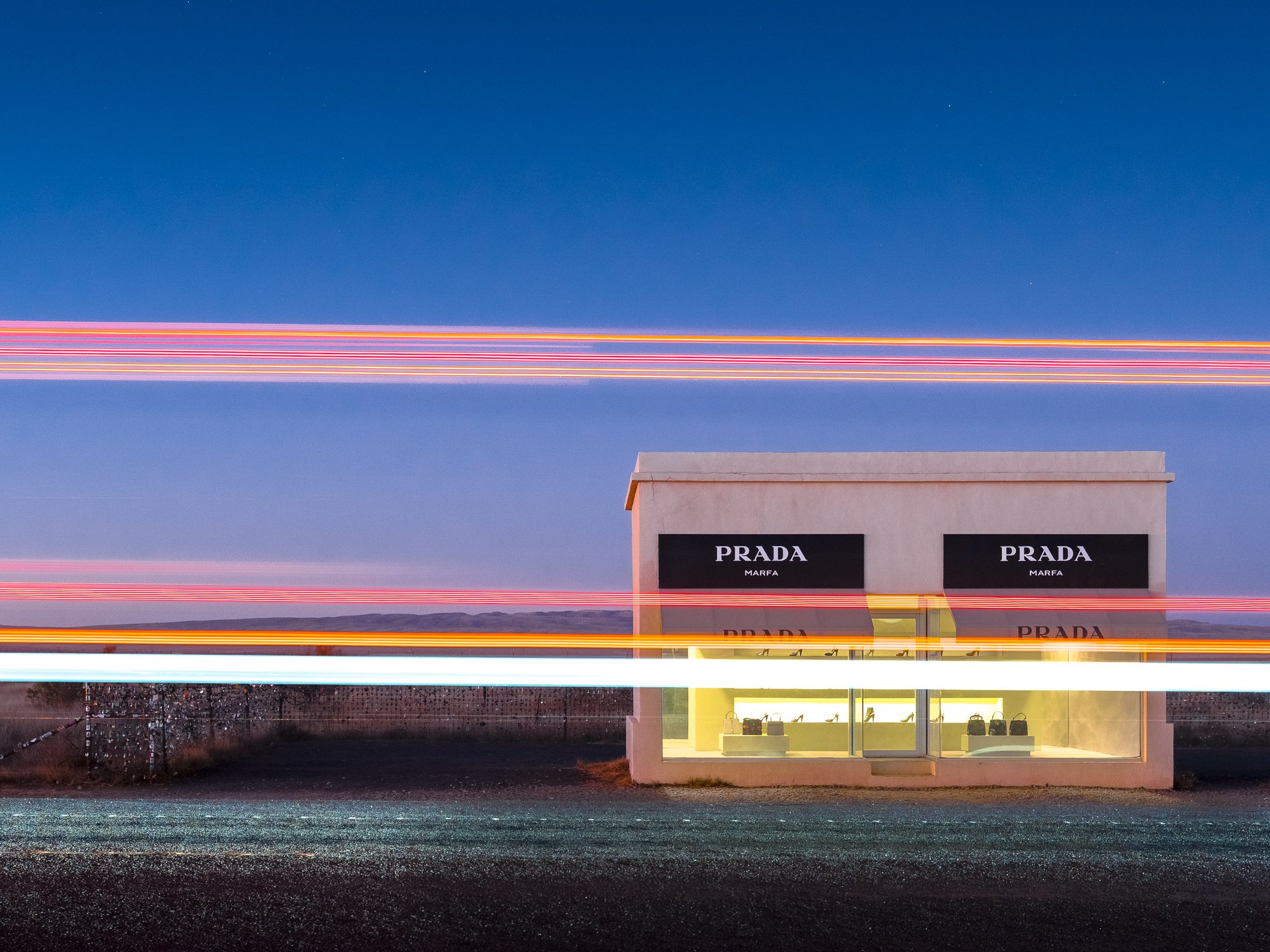 A Prada store in a desert landscape at dusk with a dark blue sky, light trails from passing vehicles, and the store's illuminated storefront with bags on display.