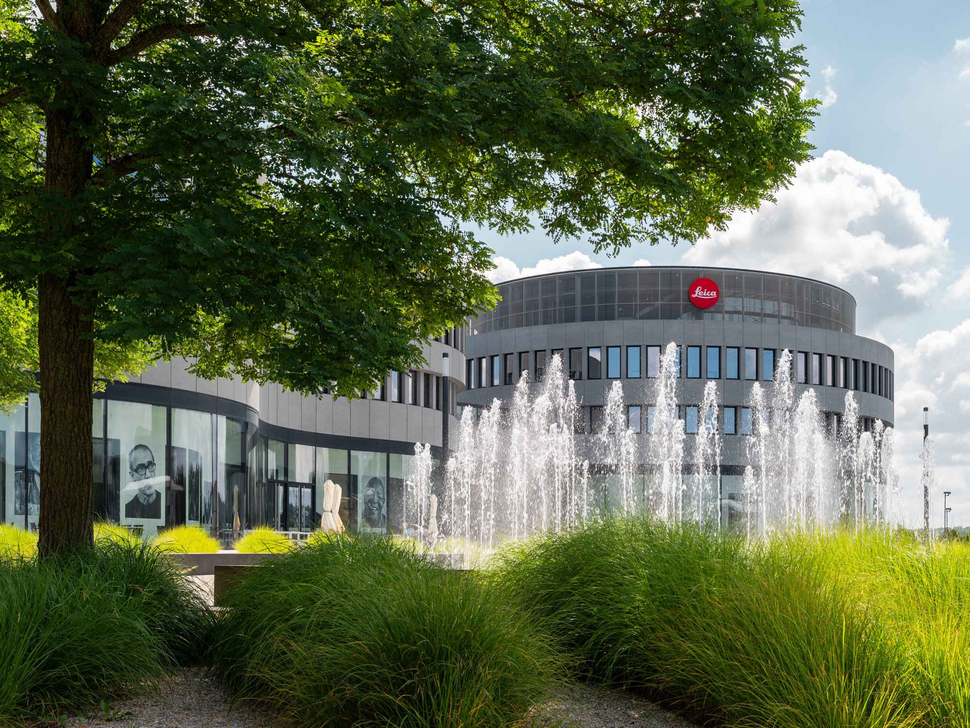 The Leica headquarters building is seen behind a fountain and green bushes, with trees and a partly cloudy sky in the background.