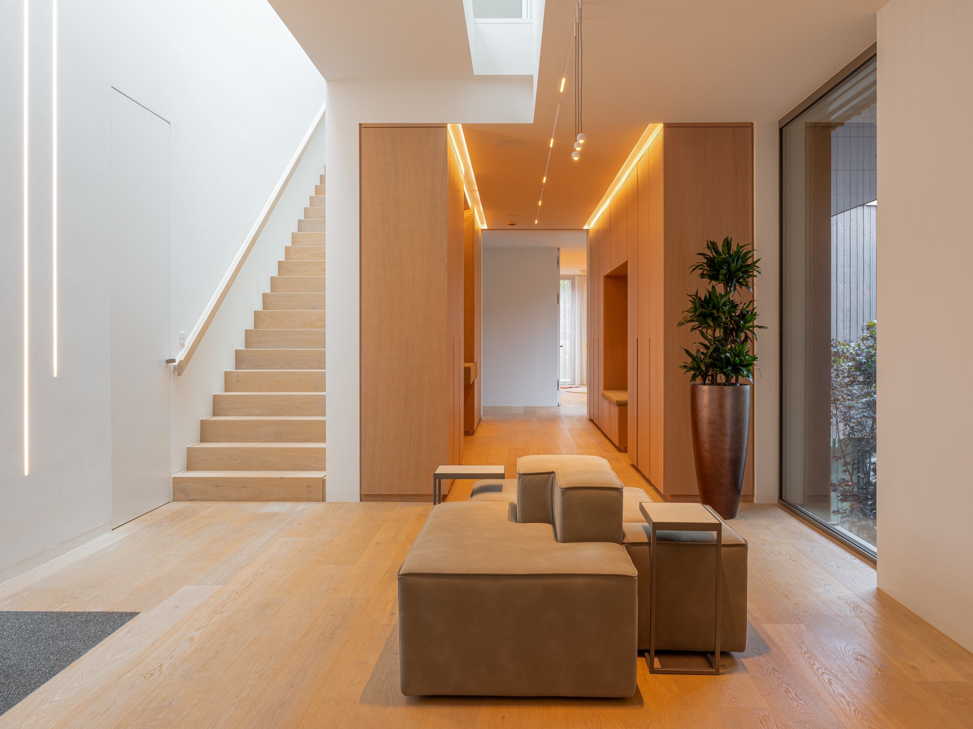 Modern interior of a house with wooden floors, a staircase on the left, a large potted plant on the right, and a cozy seating area with beige sofa and small side tables, illuminated by ceiling lights and natural light from a large window.