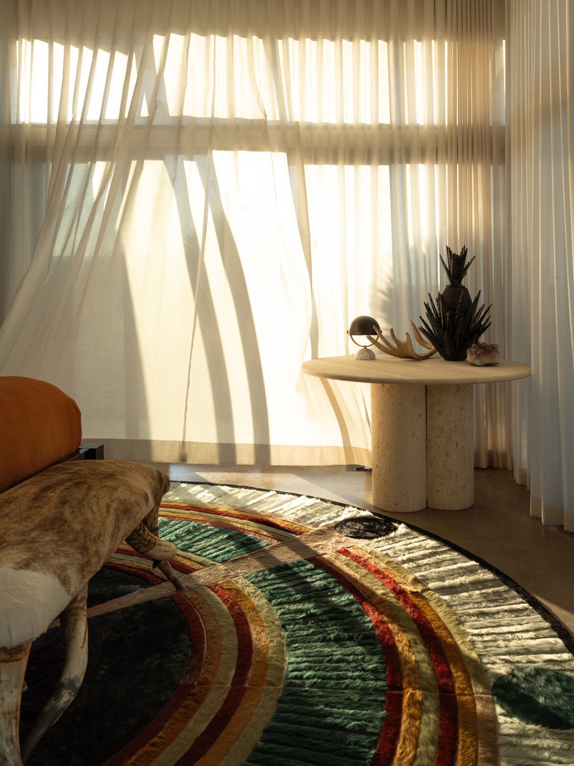 Sunlight filters through sheer curtains in a bright room with a curved multicolored rug, a wooden bench with fur and patterned cushions, and a round table holding decorative objects including a black sculpture, a shell, and a plant.