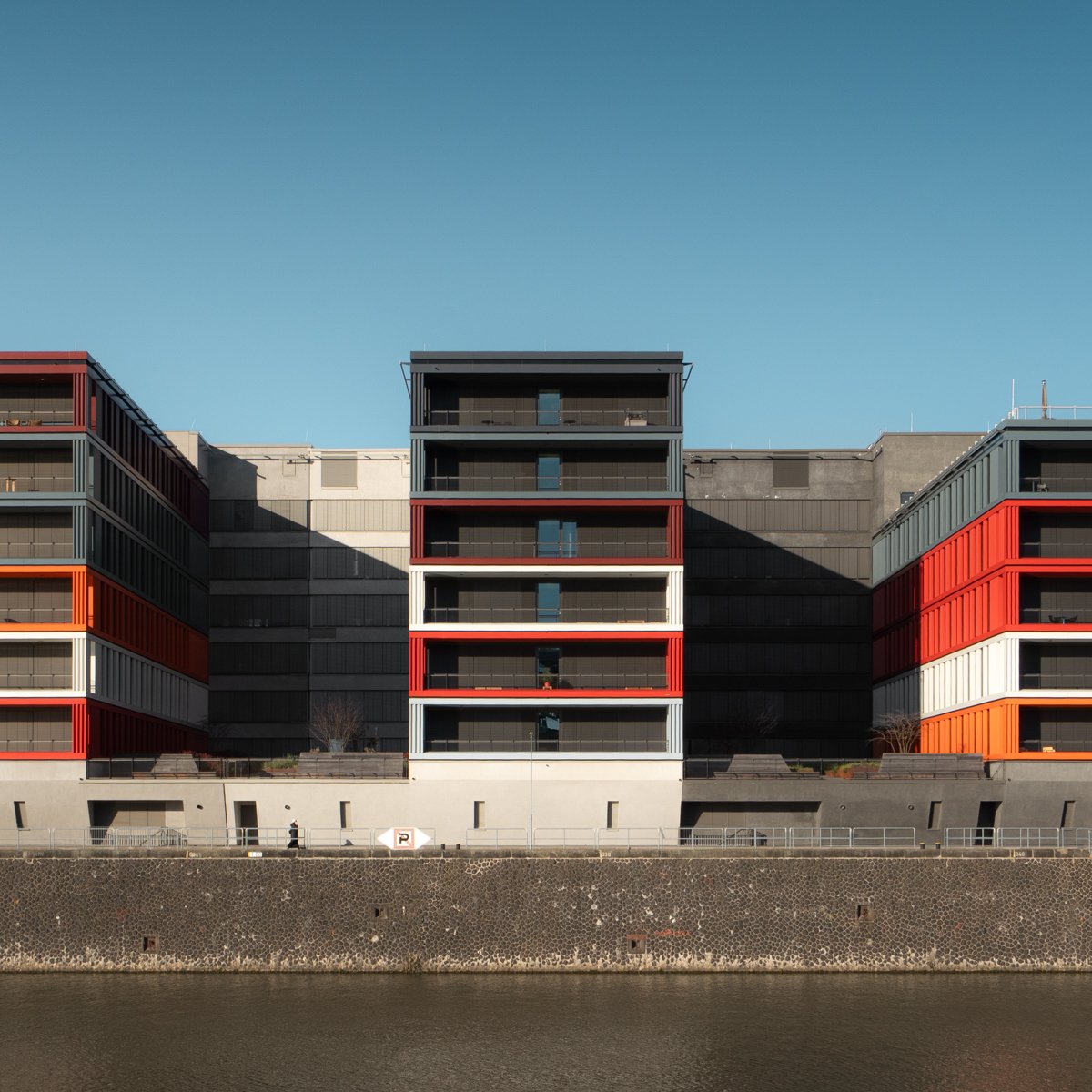 Modern apartment building with black, white, gray, and red accents, against a clear blue sky, situated by a waterway.