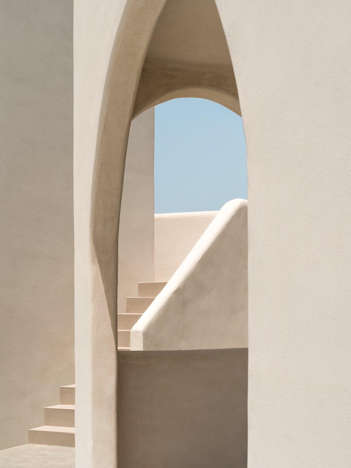 Minimalist stairway with beige walls, framed by an arched opening, against a blue sky background.