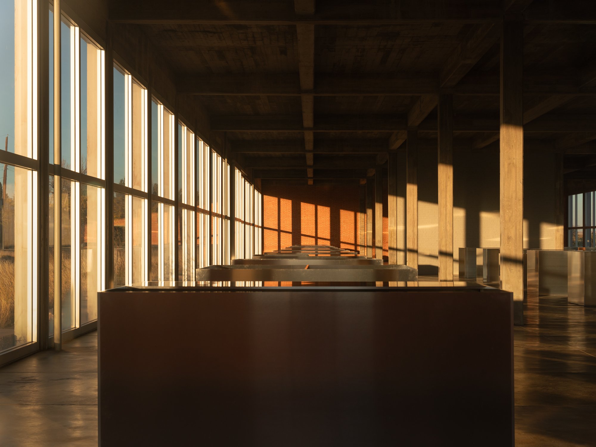 Empty interior space with large windows allowing sunlight to cast shadows inside a modern building with wooden beams and concrete supports.