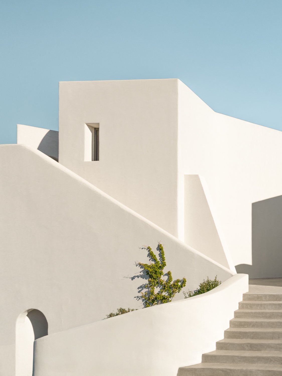Modern white building with geometric shapes, steps, and minimal landscaping against a clear blue sky.