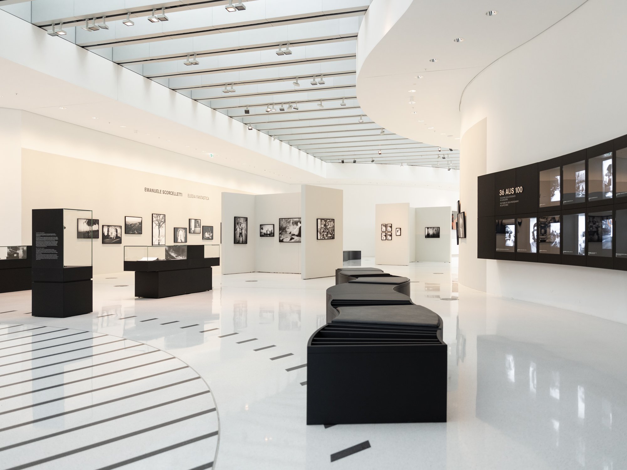 Modern art gallery interior with white walls, black display cases, and black and white photographs on the walls. Curved seating and exhibits are visible, with a glass ceiling allowing natural light.