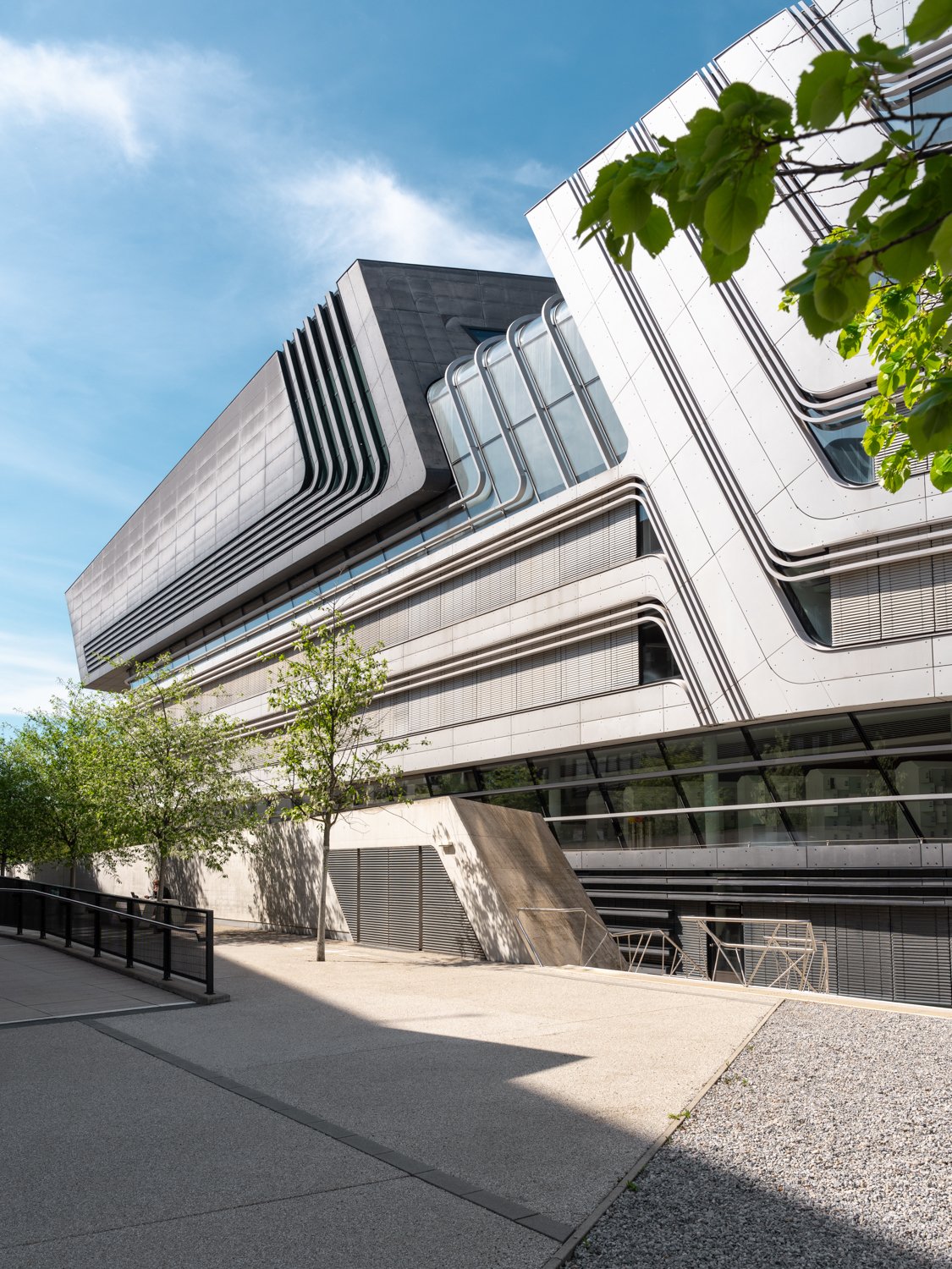 Modern multi-story building with sleek architectural design, metal and glass exterior, trees in foreground, and blue sky with clouds.