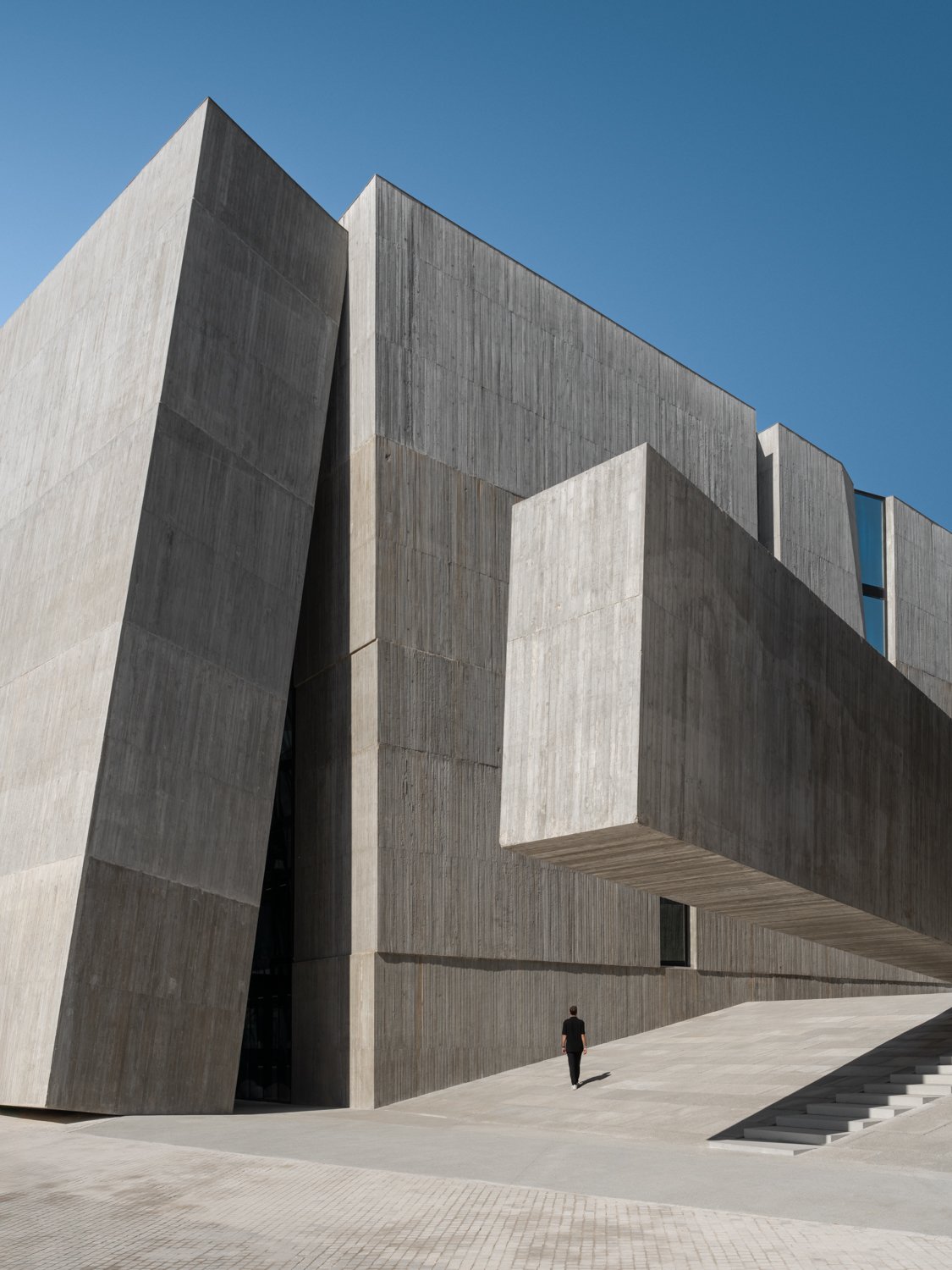A person walking near a large modern concrete building with sharp angles and geometric shapes against a clear blue sky.