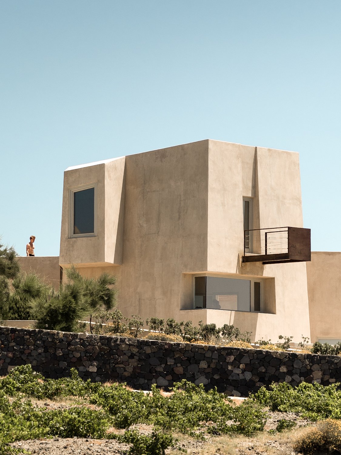 Modern beige stucco house with angular design, large windows, and a small balcony, surrounded by desert plants and rocky landscaping under a clear blue sky.