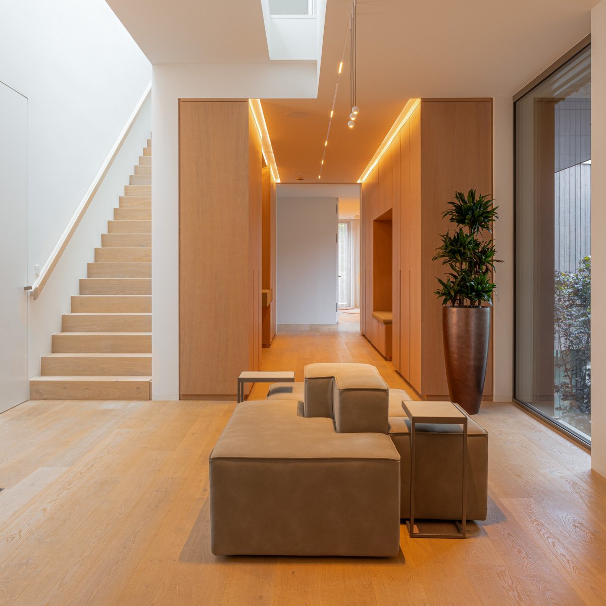 Modern interior with wooden floors, staircase, large window, beige seating, and tall potted plant.