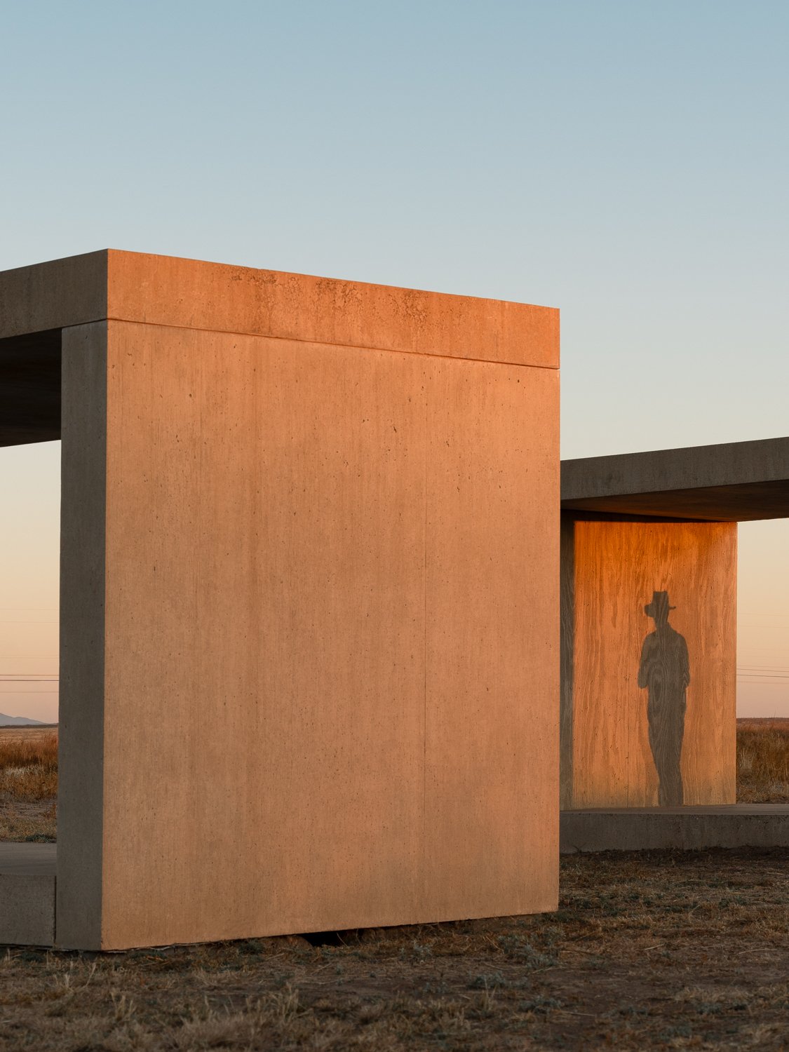 Concrete modern structure with rectangular shapes in a field during sunset, featuring the shadow of a person wearing a hat projected onto the wall.