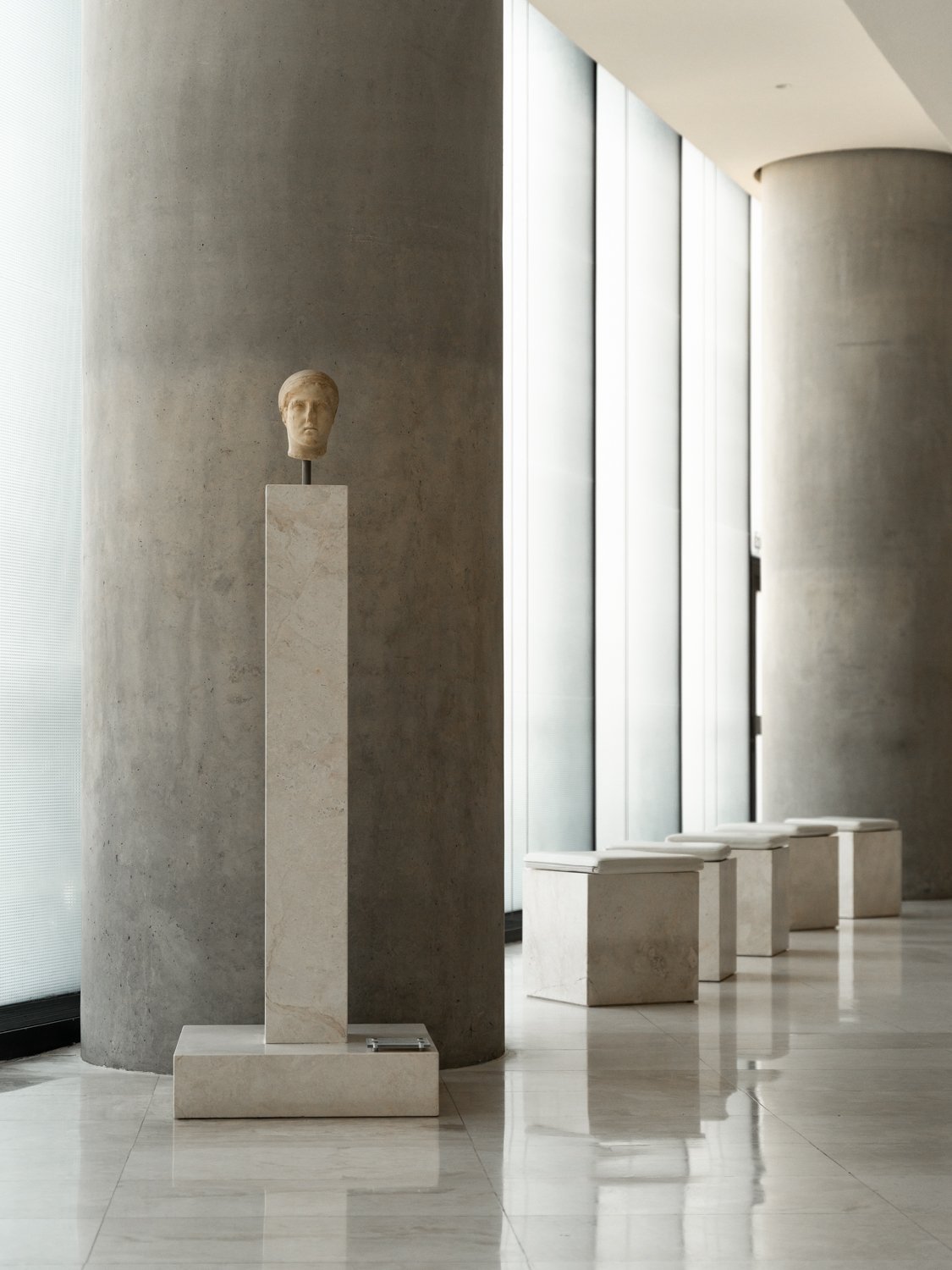 Modern interior with a marble bust on a pedestal, beige benches, large windows, and concrete walls.