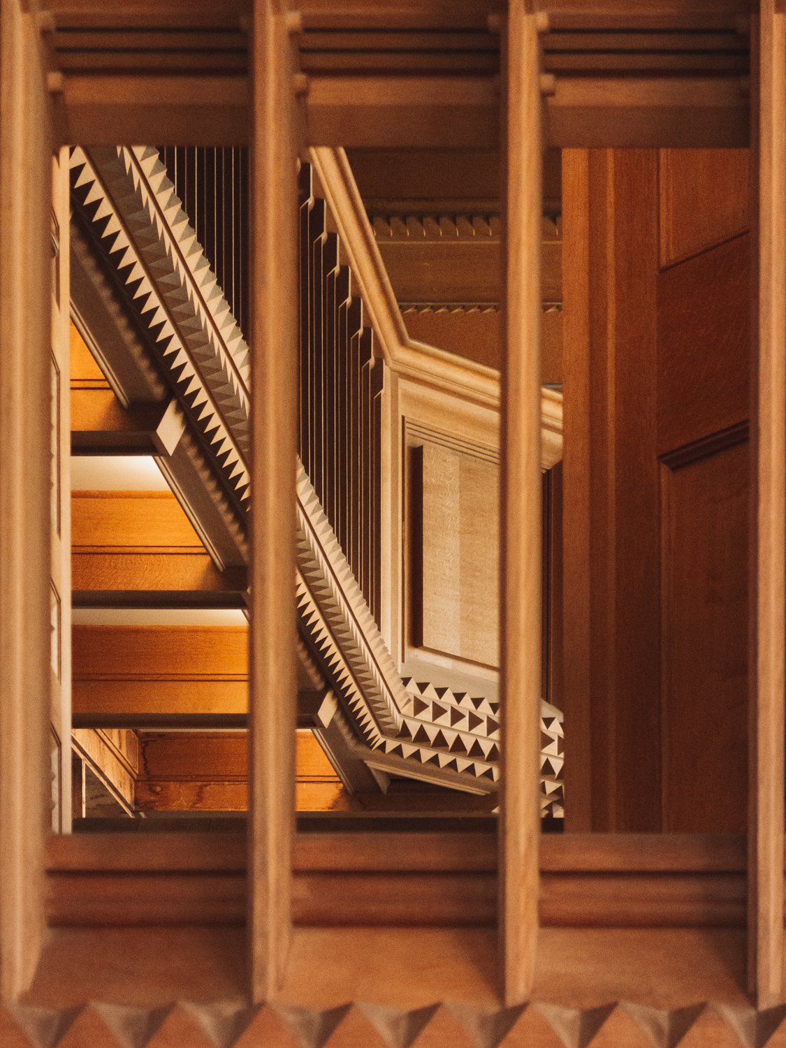 View of a multi-level wooden staircase through vertical wooden railing bars, showing the patterned edges of each staircase.
